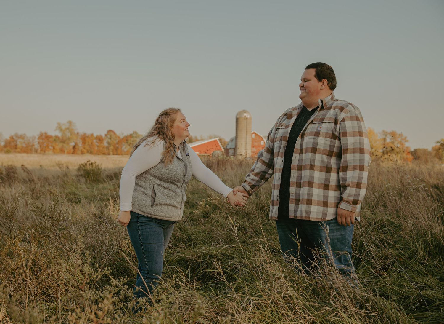 Couple standing together in a field during sunset near a barn.