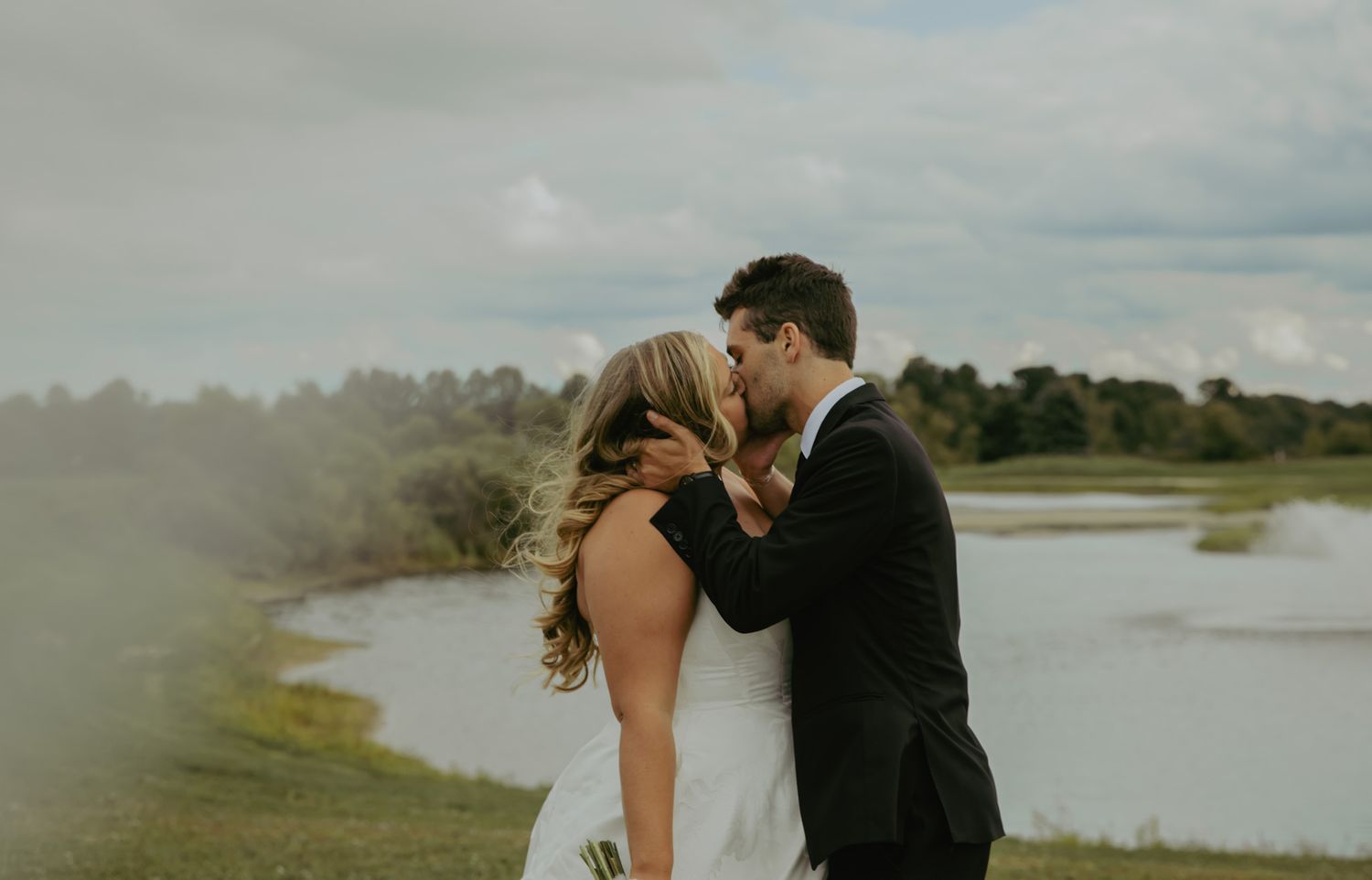 A bride and groom share a romantic kiss outdoors near a scenic lake on their wedding day.