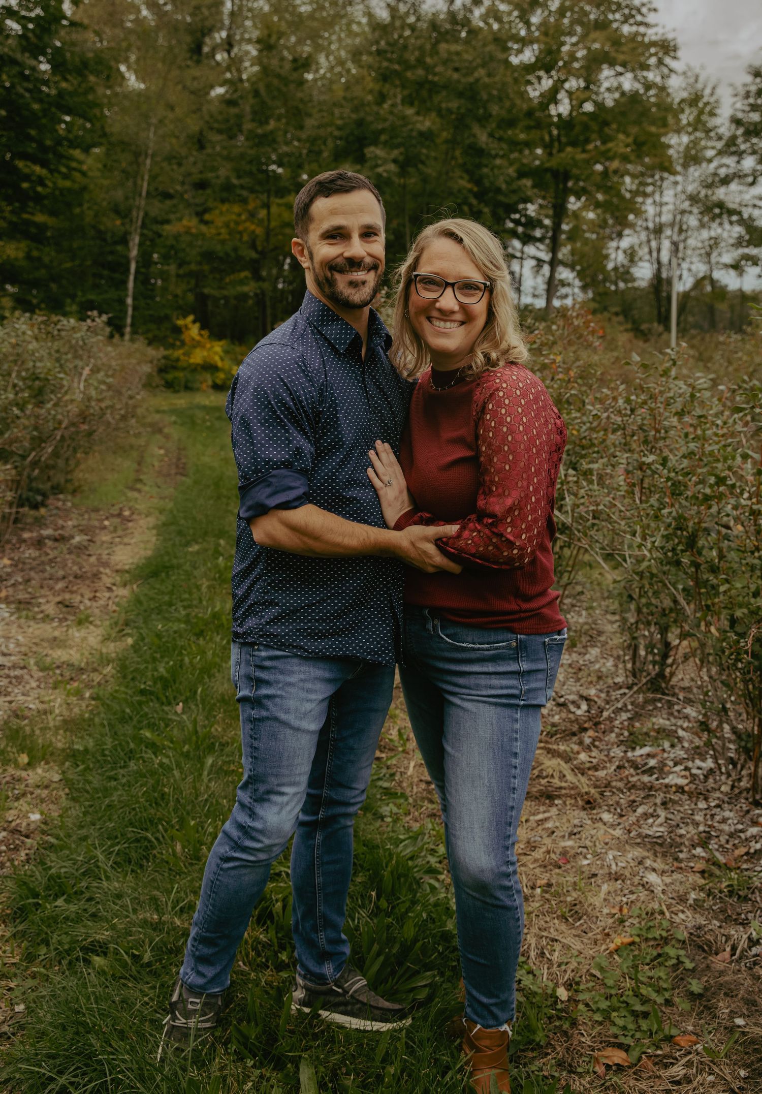 Smiling couple embracing outdoors on a garden path surrounded by lush green trees in autumn.