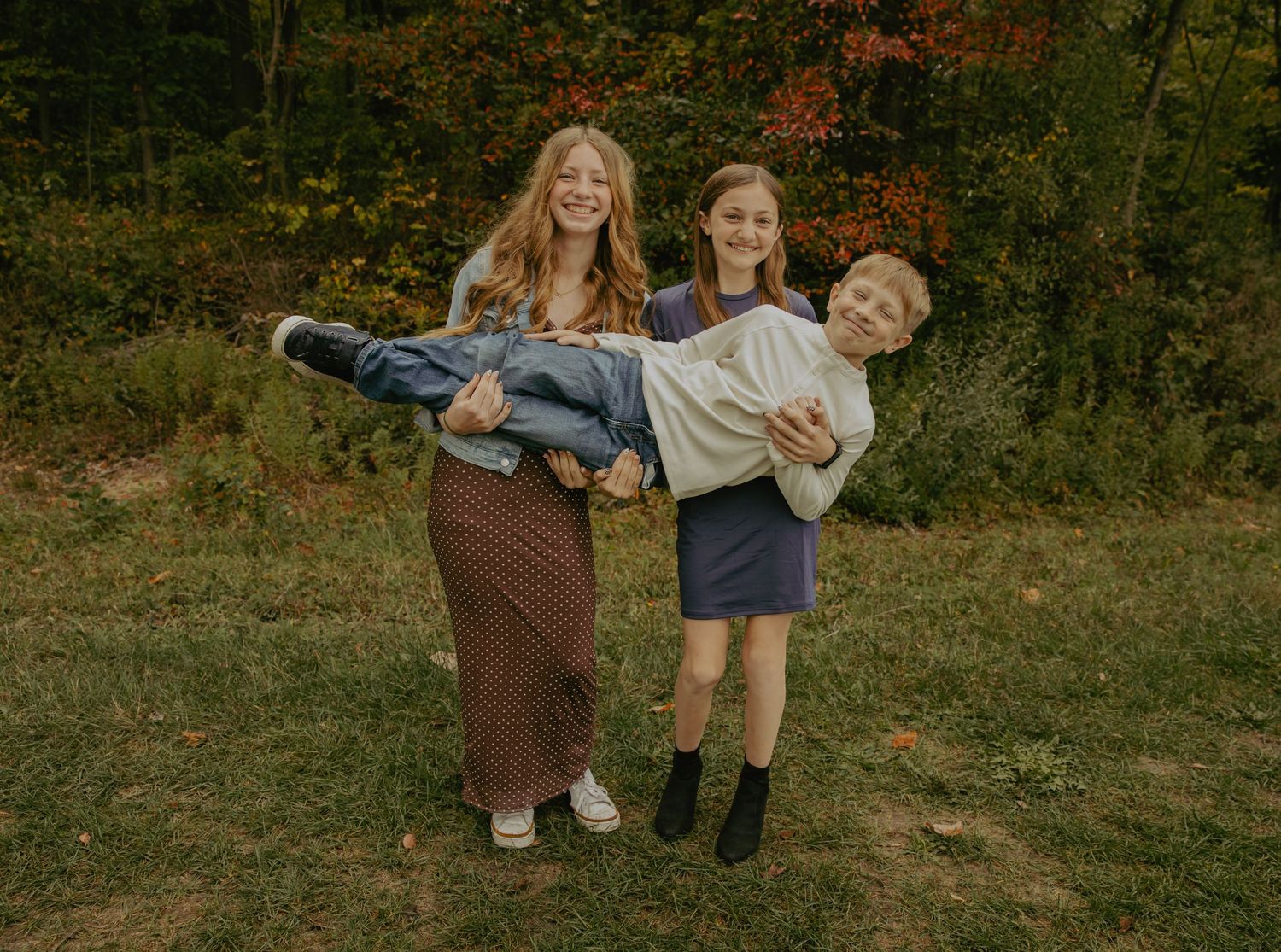 Three siblings pose playfully outdoors in fall, two girls carrying a boy, surrounded by colorful autumn foliage.