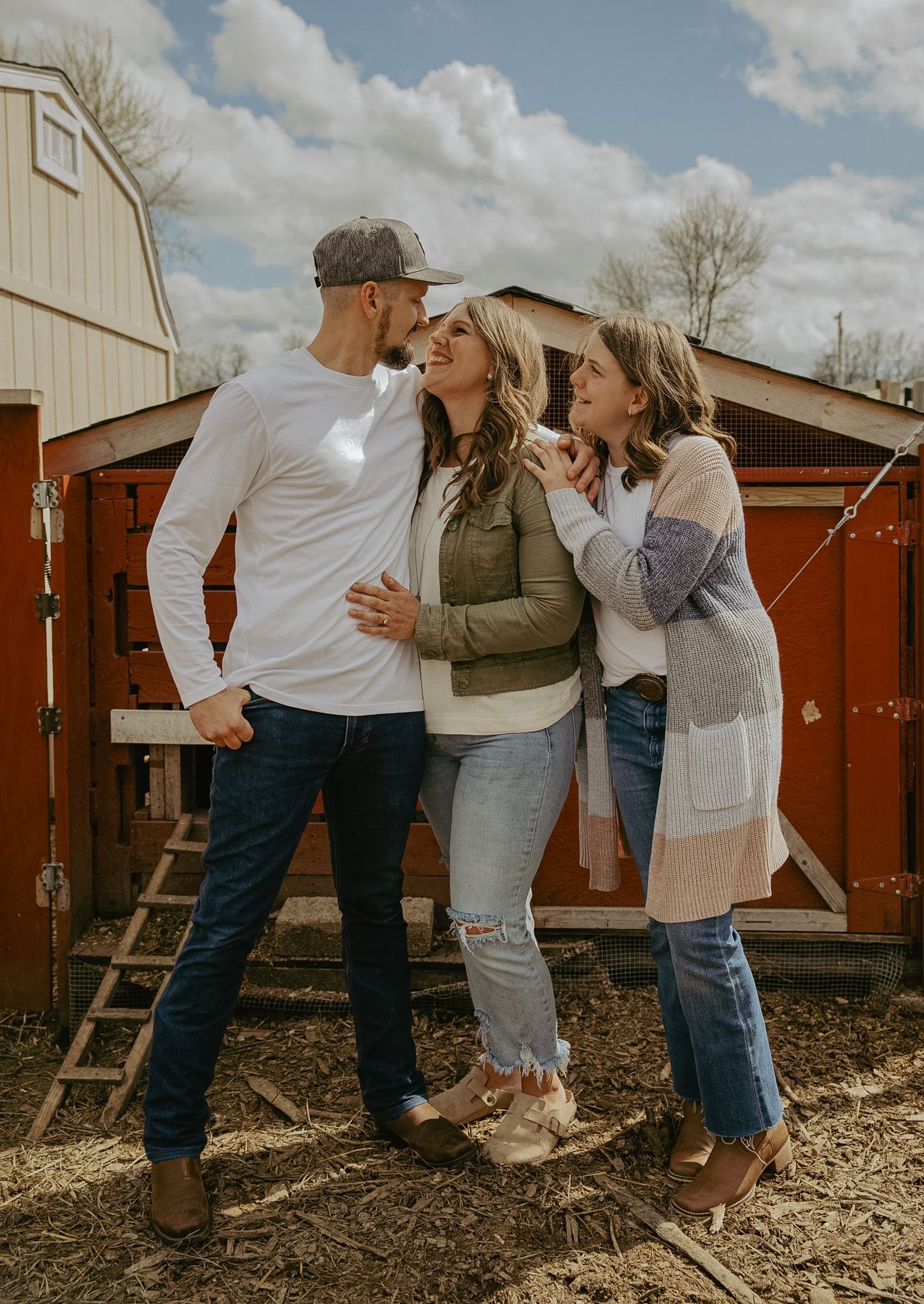 Three friends smiling and embracing outdoors near rustic red barn on a sunny day.