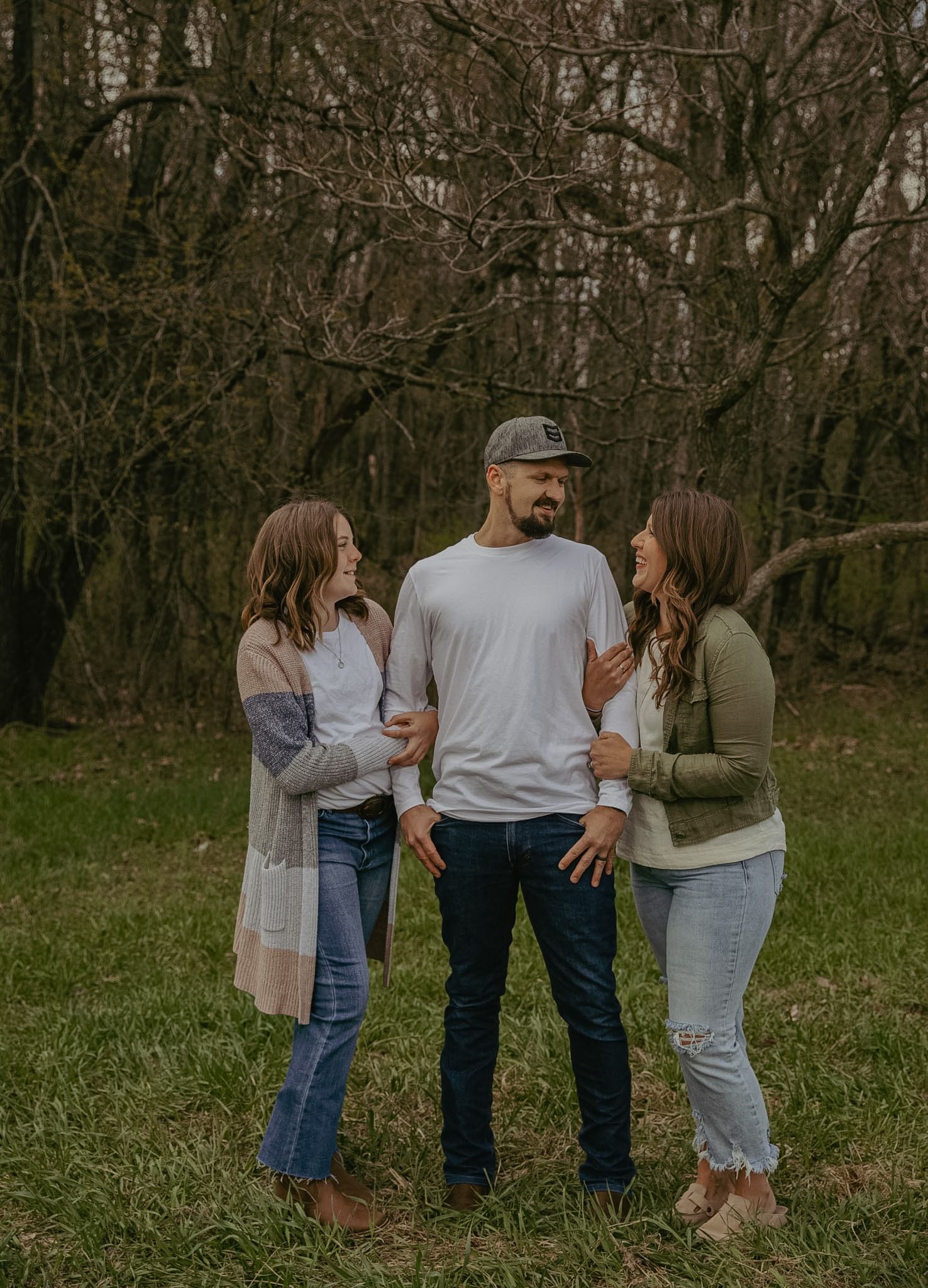 A man in a white shirt and cap stands between two women in casual outfits, smiling outdoors near bare trees.