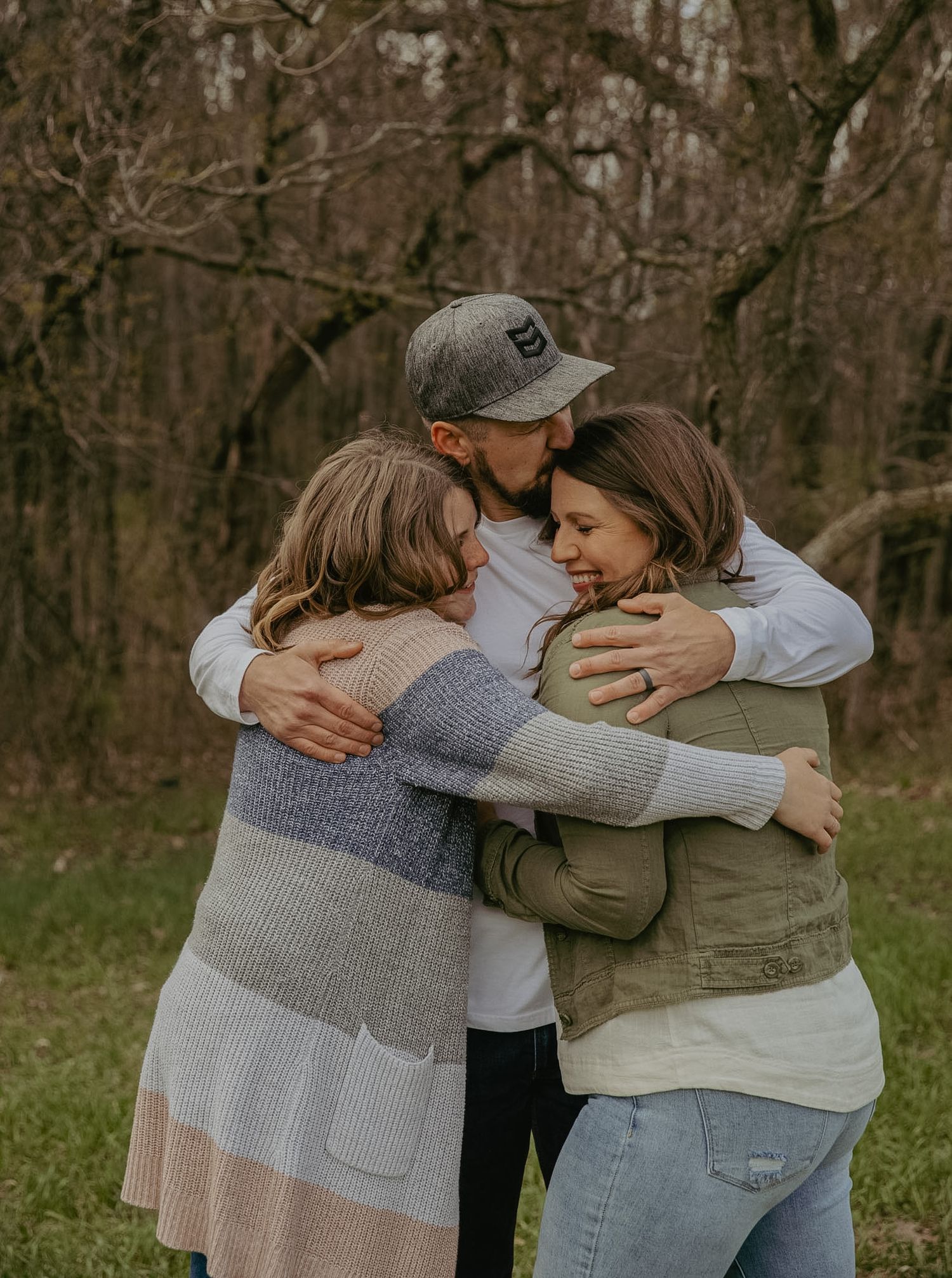 Three people share a warm group hug outdoors in a wooded area during a family photo session.
