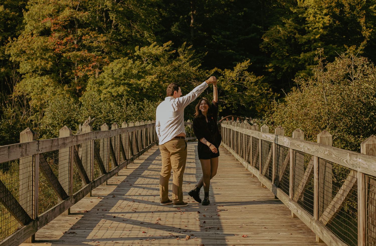 Couple walks along wooden bridge pathway surrounded by autumn foliage.
