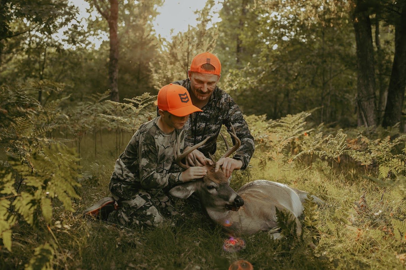 A hunter in camouflage gear and orange cap kneels beside a harvested whitetail deer in an autumn forest setting.