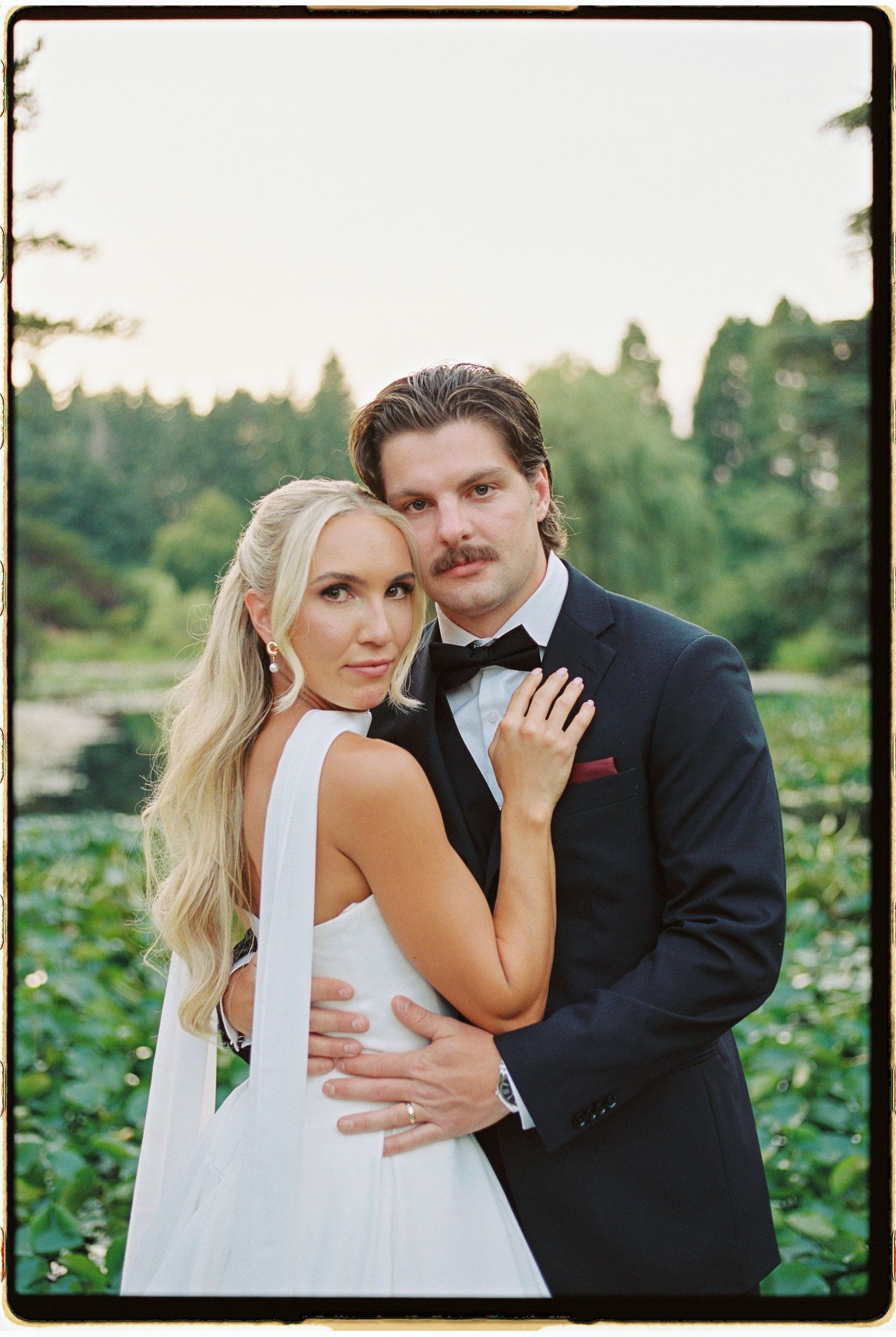 Elegant couple in wedding attire embrace outdoors, groom in navy tuxedo, bride in white gown, lush green trees behind.