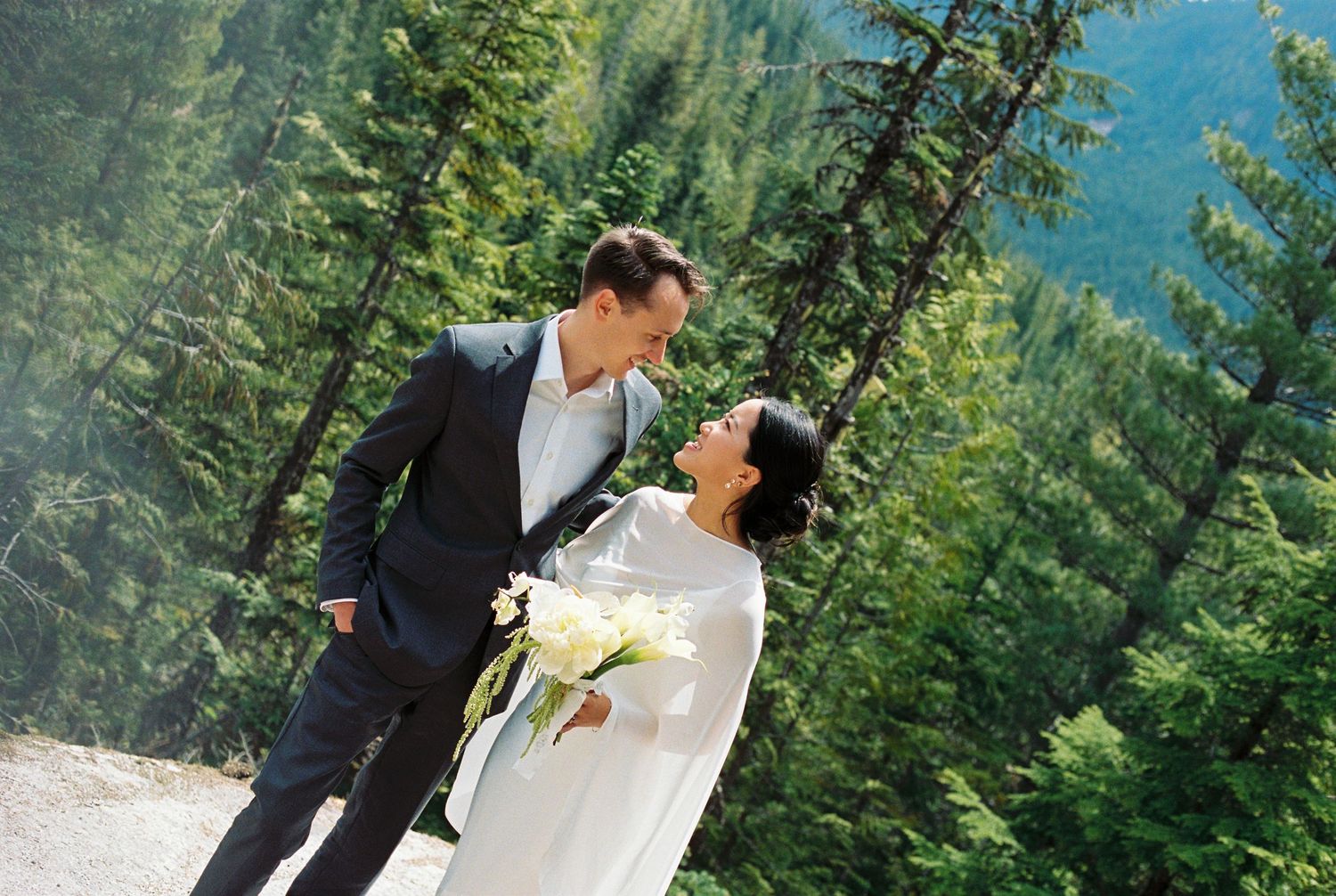 Bride and groom sharing a romantic moment outdoors with lush green pine trees and mountain scenery in the background.