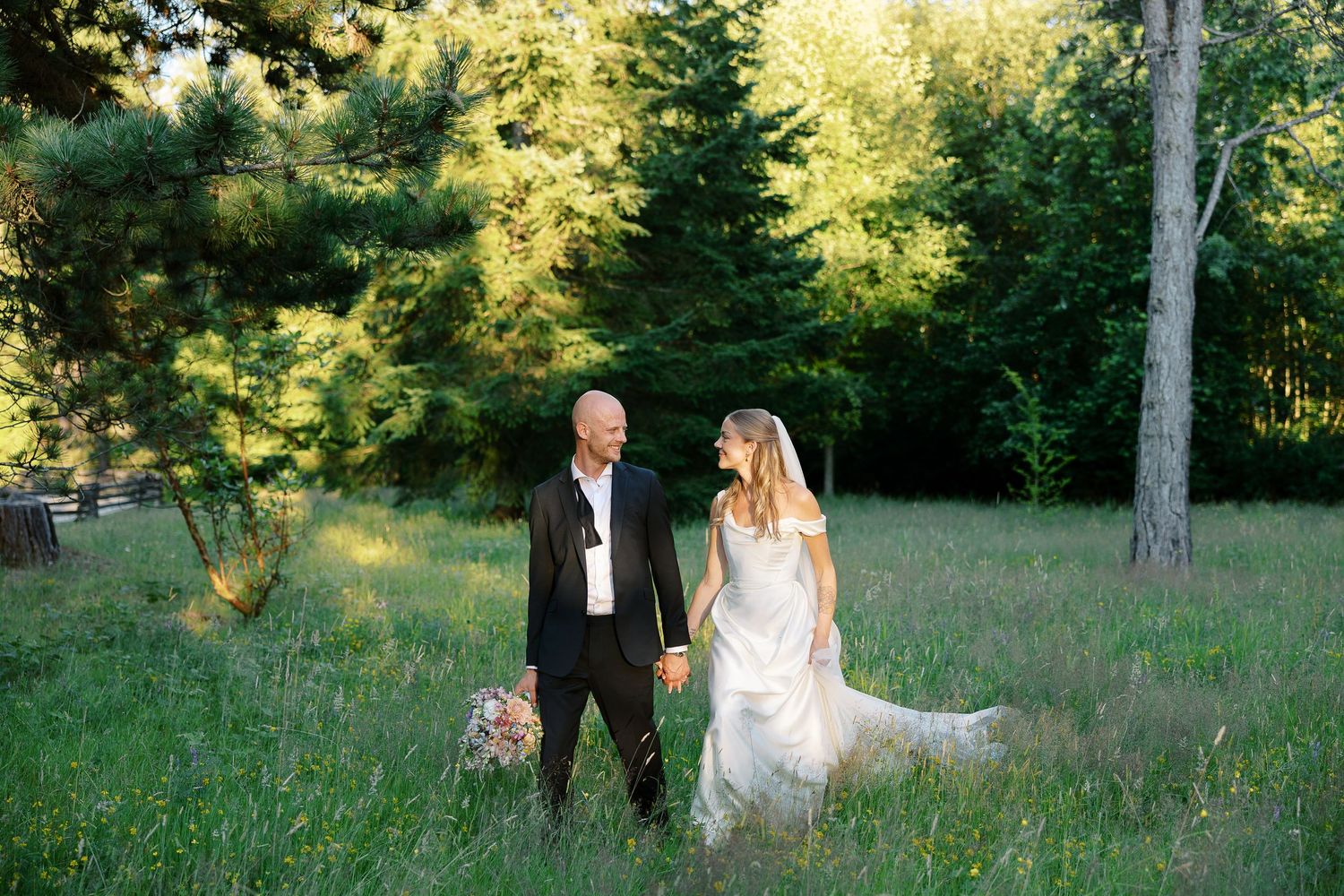 Bride and groom holding hands in a lush green meadow surrounded by tall trees during golden hour outdoor wedding.