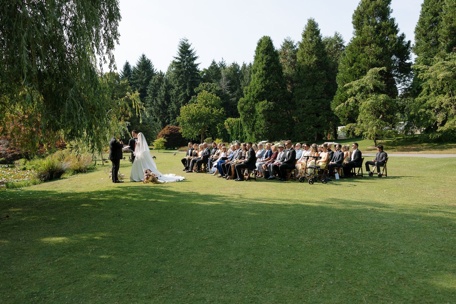 Outdoor wedding ceremony on lush green lawn surrounded by tall trees with guests seated in rows facing the couple.