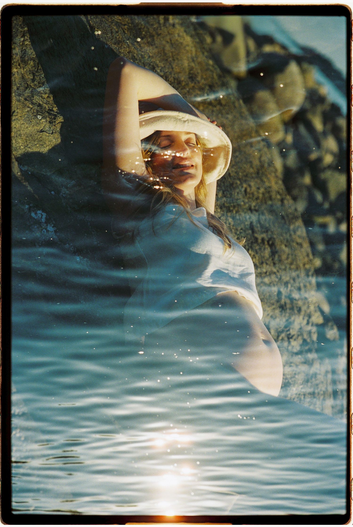 Woman in white hat relaxing against rocks by shimmering water in golden sunlight, dreamy double exposure photography.