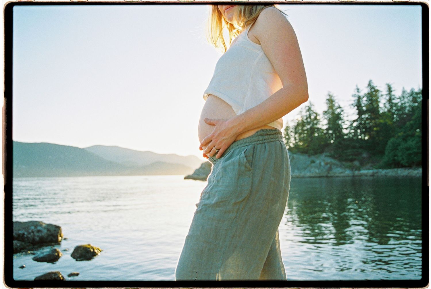 Pregnant woman in white crop top and green pants cradling her baby bump by a serene lake at sunset.
