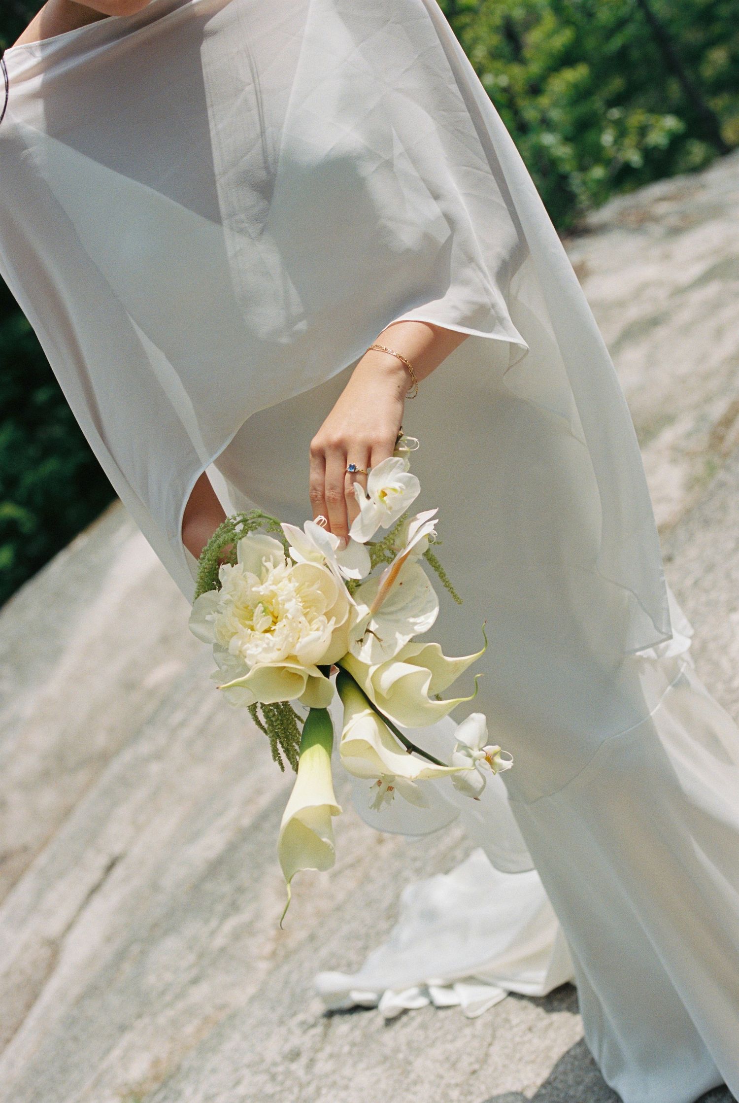 Bride in flowing white gown holding elegant bouquet of white calla lilies and peonies outdoors.