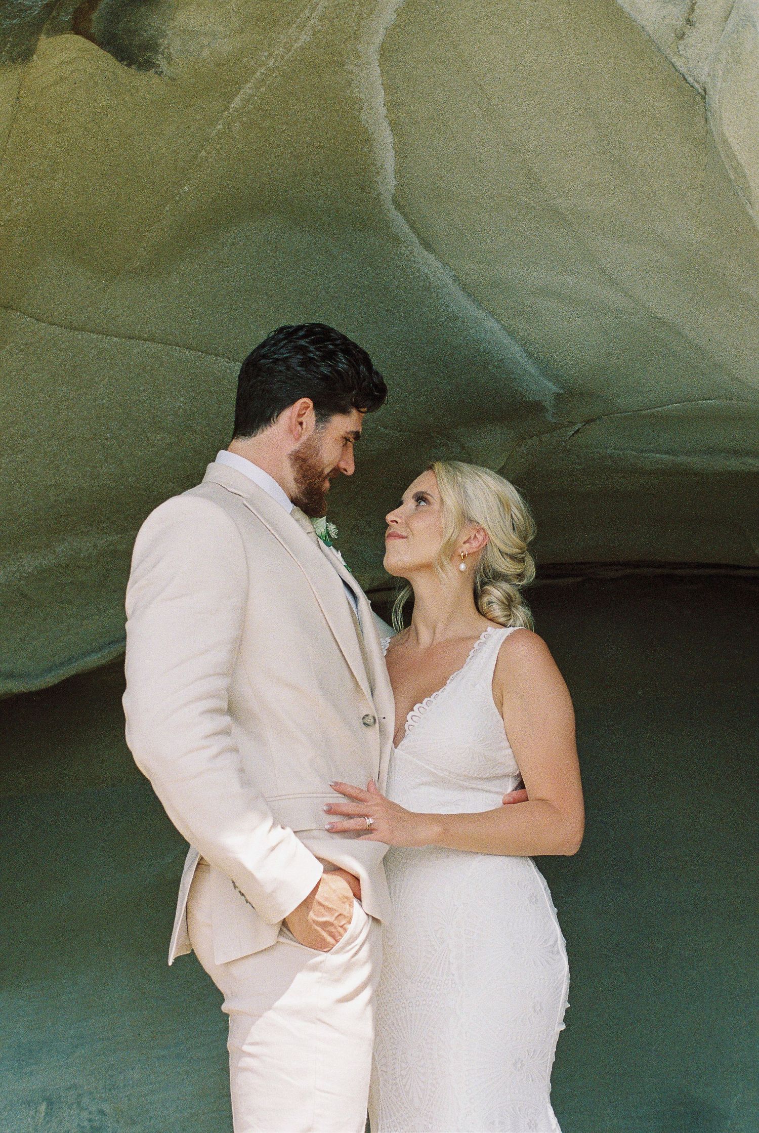 A bride and groom share an intimate moment in a sandy cave, she in a white gown, he in a beige suit.