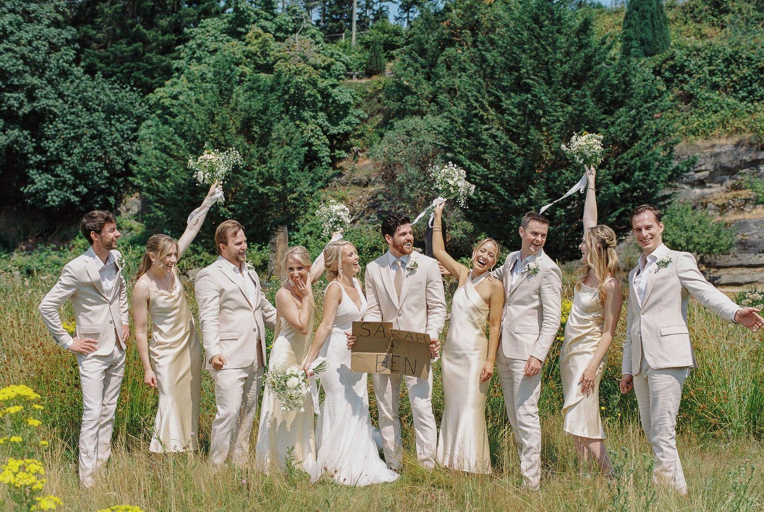 Wedding party celebrating outdoors in a meadow, bridesmaids in gold dresses and groomsmen in white suits raising bouquets.