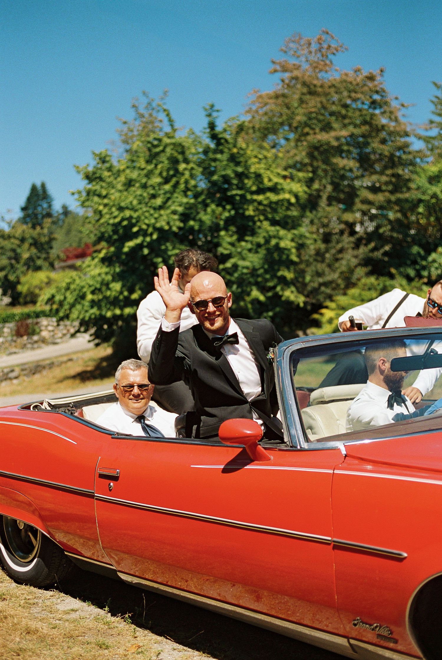 Groom waving from a classic red convertible car on a sunny day with trees in the background.