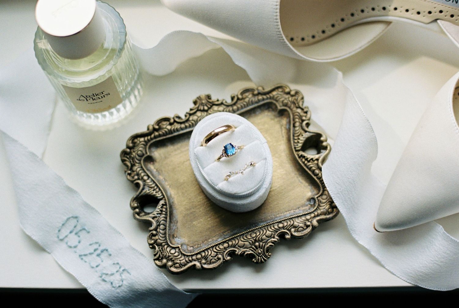 Elegant wedding flat lay with gold ornate frame, blue gemstone engagement ring, wedding band, perfume bottle, and white ribbon.