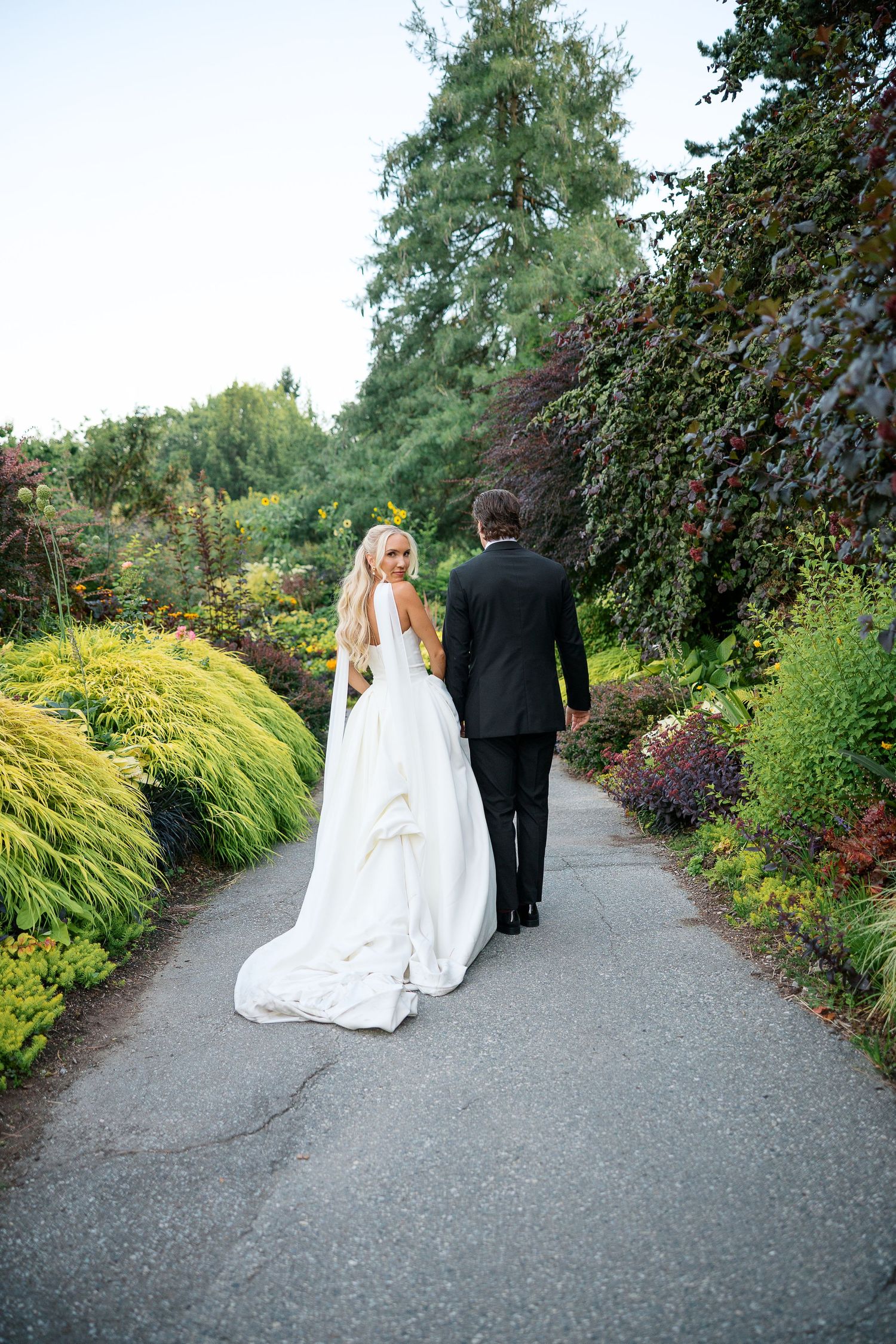 Bride and groom walking hand in hand down a lush garden path, bride wearing a flowing white gown with long train.