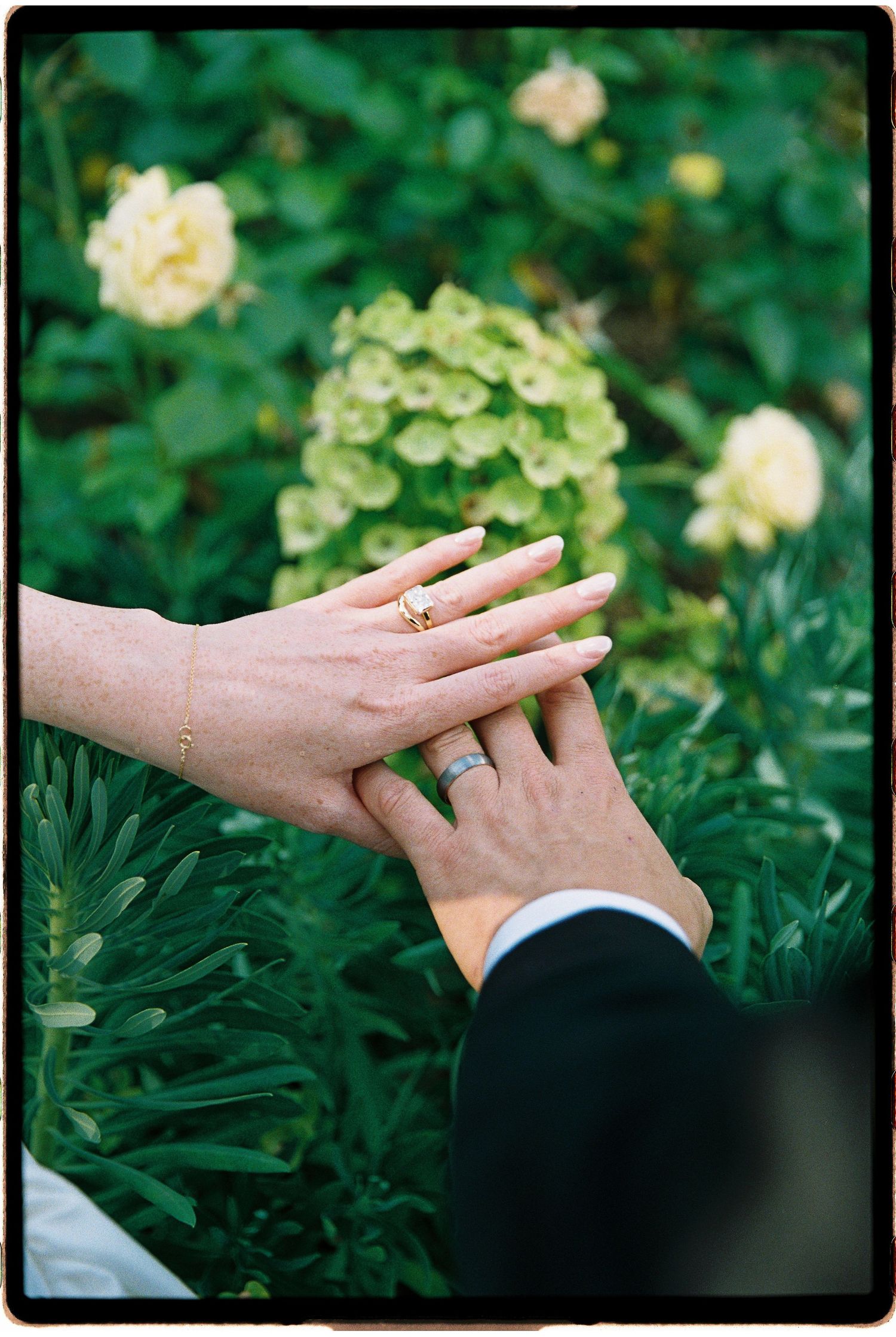 Newlywed couple holding hands showing wedding rings in a lush garden with yellow roses and green hydrangeas.