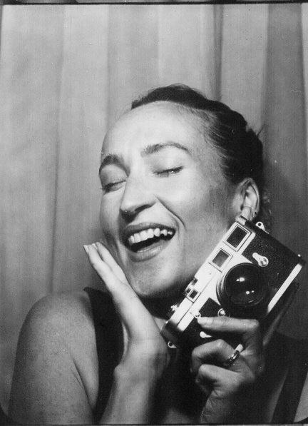 Black and white photo of a laughing woman holding a vintage Leica camera, eyes closed, hand on cheek.