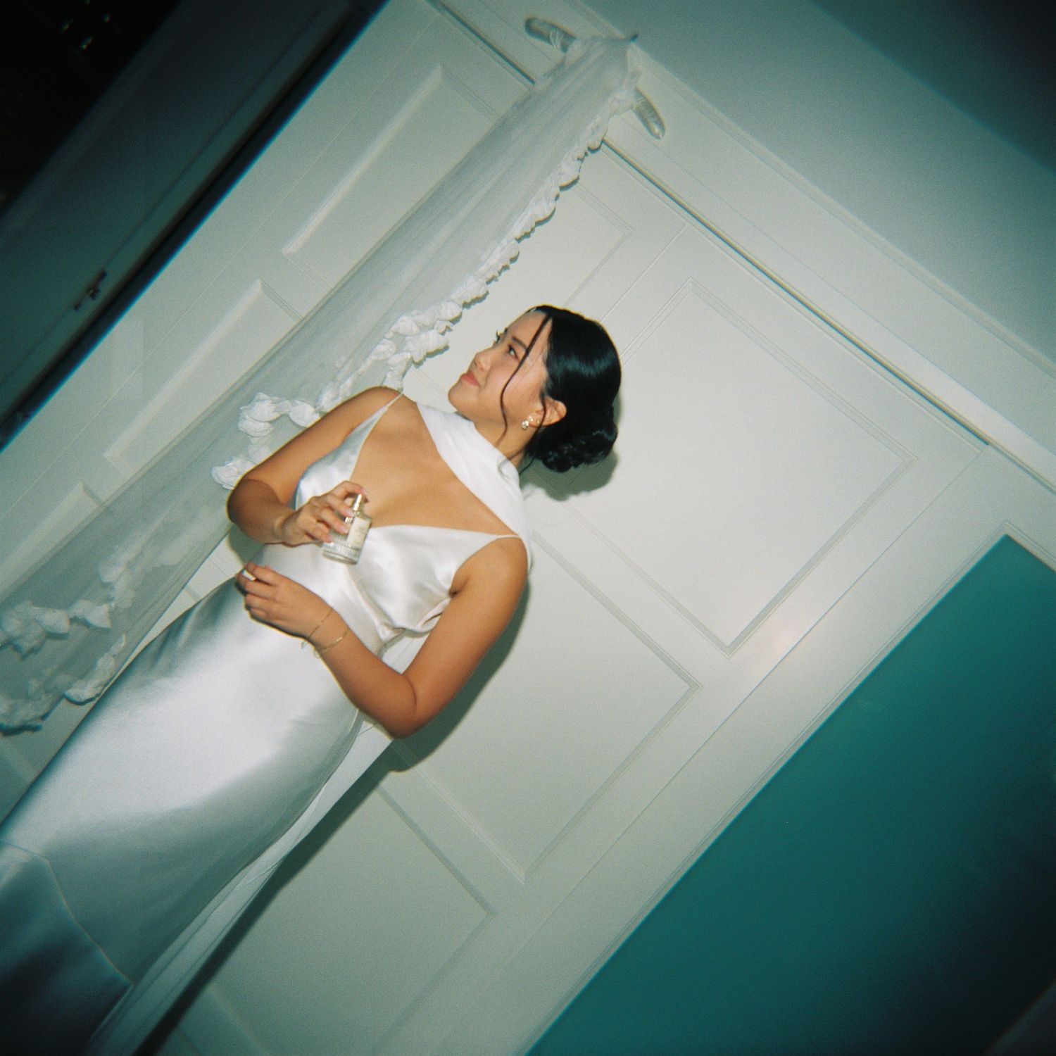 A woman in a white satin dress holds a glass near a white door in a softly lit room.