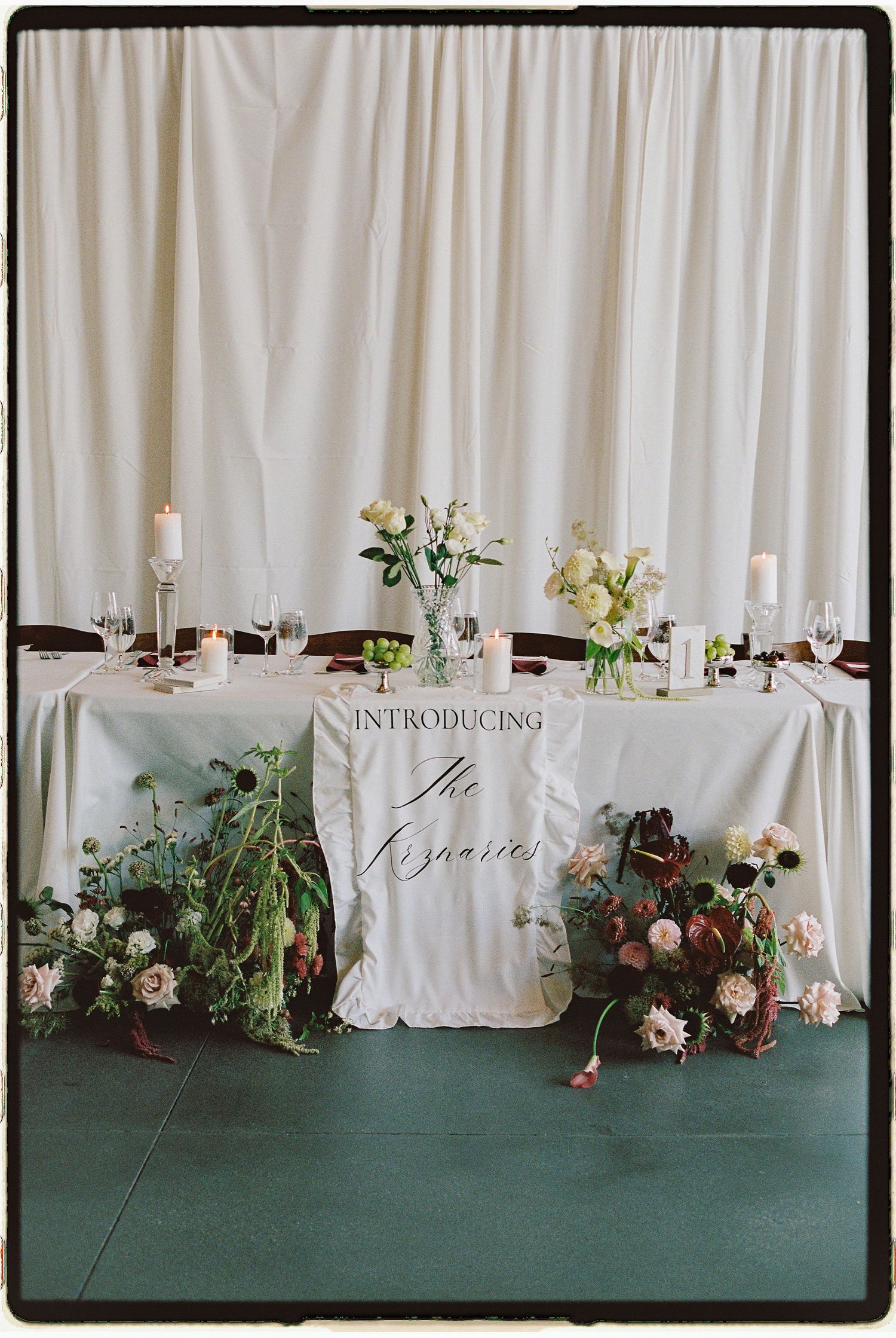 Elegant wedding sweetheart table with white linen, candles, floral arrangements, and an introductory newlyweds sign.