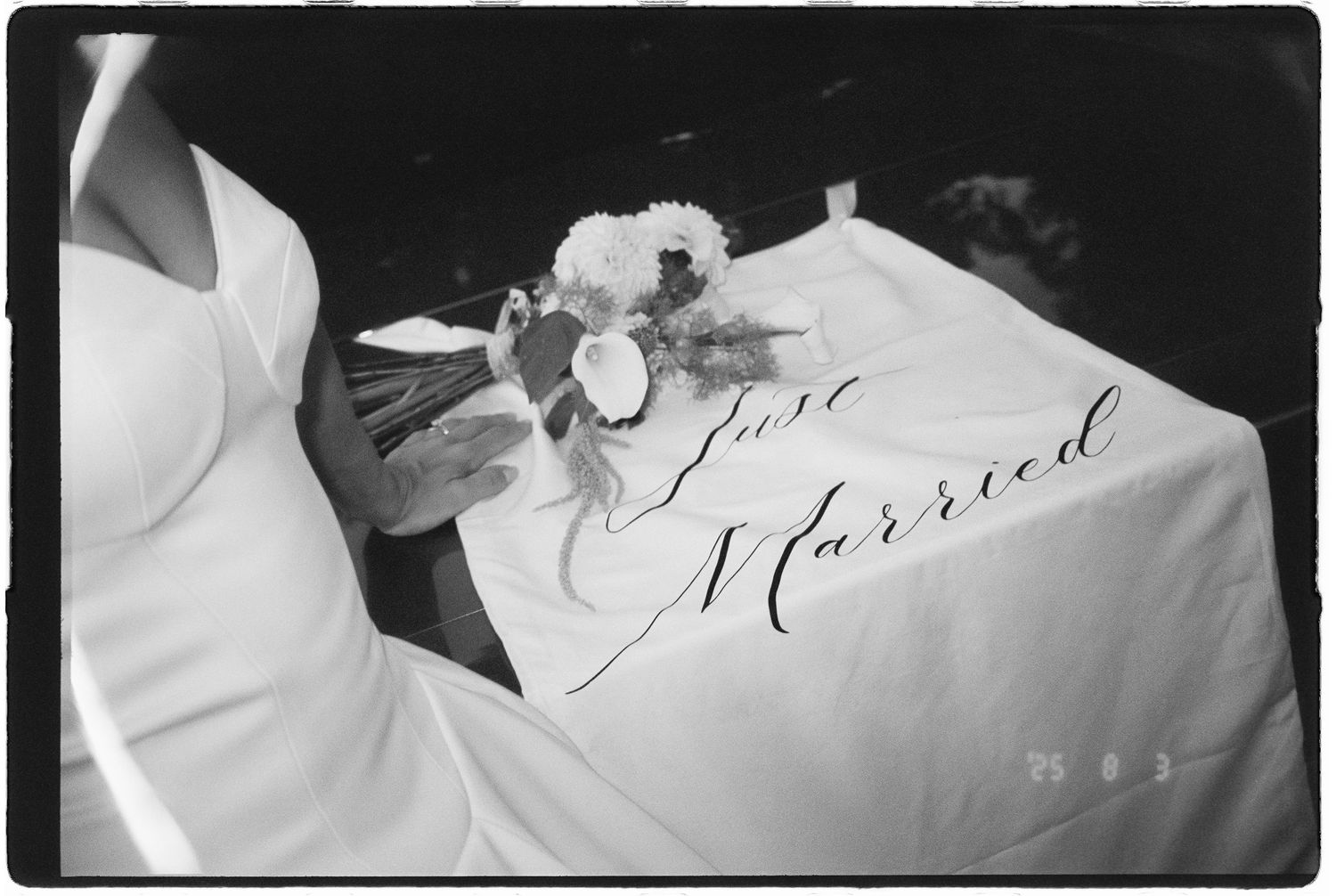 Black and white photo of a Just Married sign on a white tablecloth with a bridal bouquet beside a wedding dress.