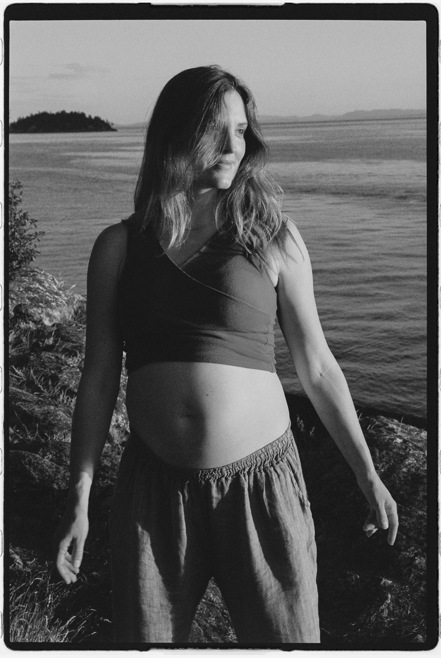Black and white photo of a young woman with long hair standing outdoors near water, wearing a crop top and skirt.
