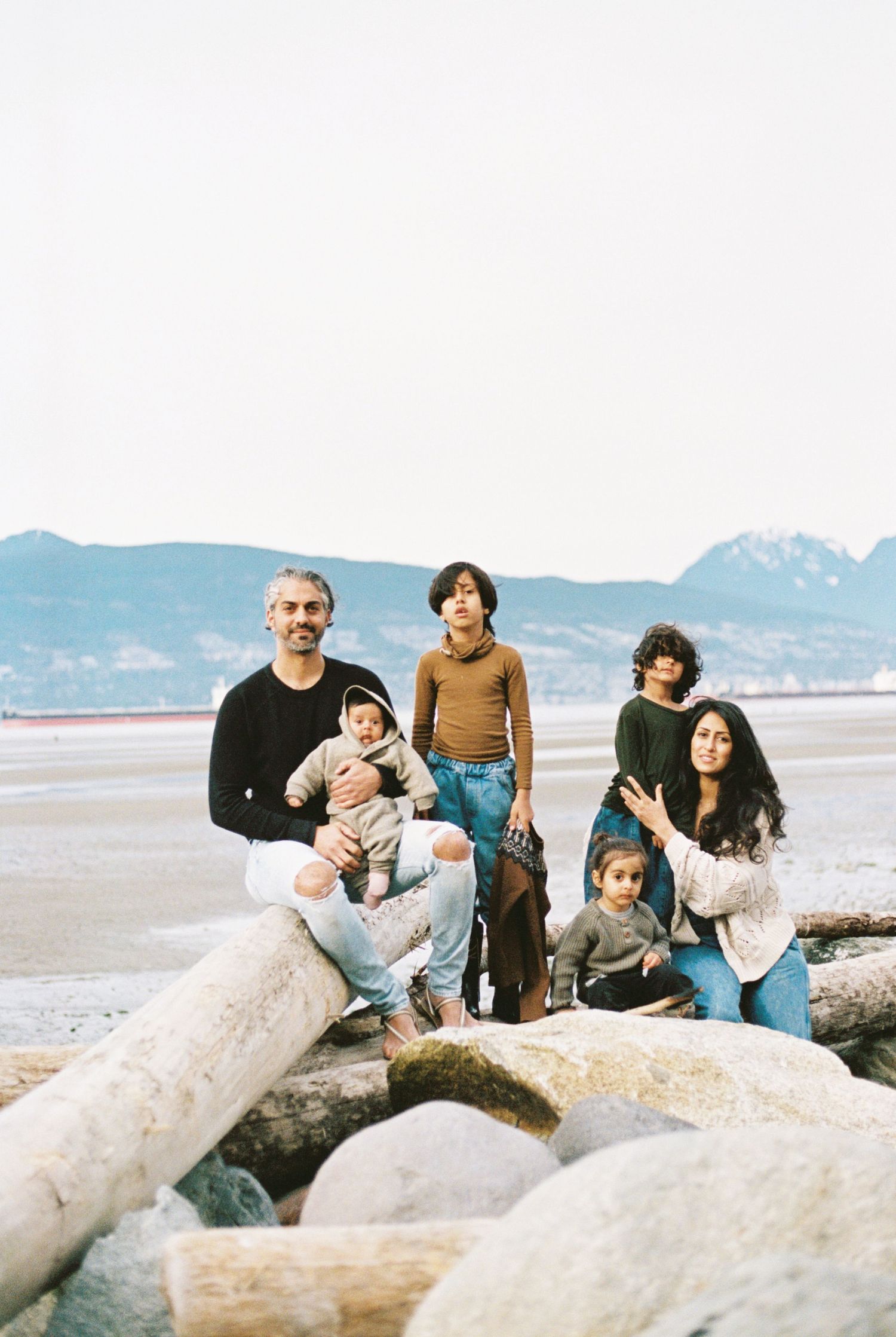 Group sitting together on driftwood logs at a beach with mountains in the background.