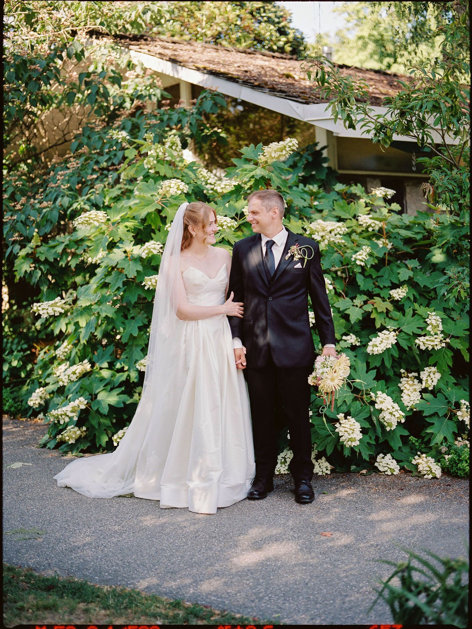 Bride in white gown and groom in black tuxedo stand together smiling amid lush green flowering bushes outdoors.