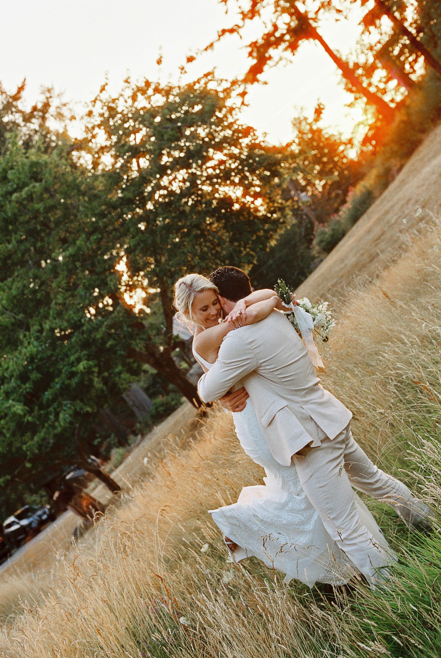 A joyful wedding couple spins and embraces in a golden field at sunset.
