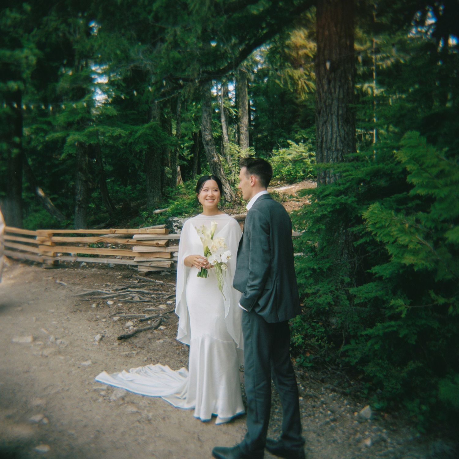 A bride and groom pose on a forest path, she in a white gown holding flowers, he in a gray suit, surrounded by tall trees.
