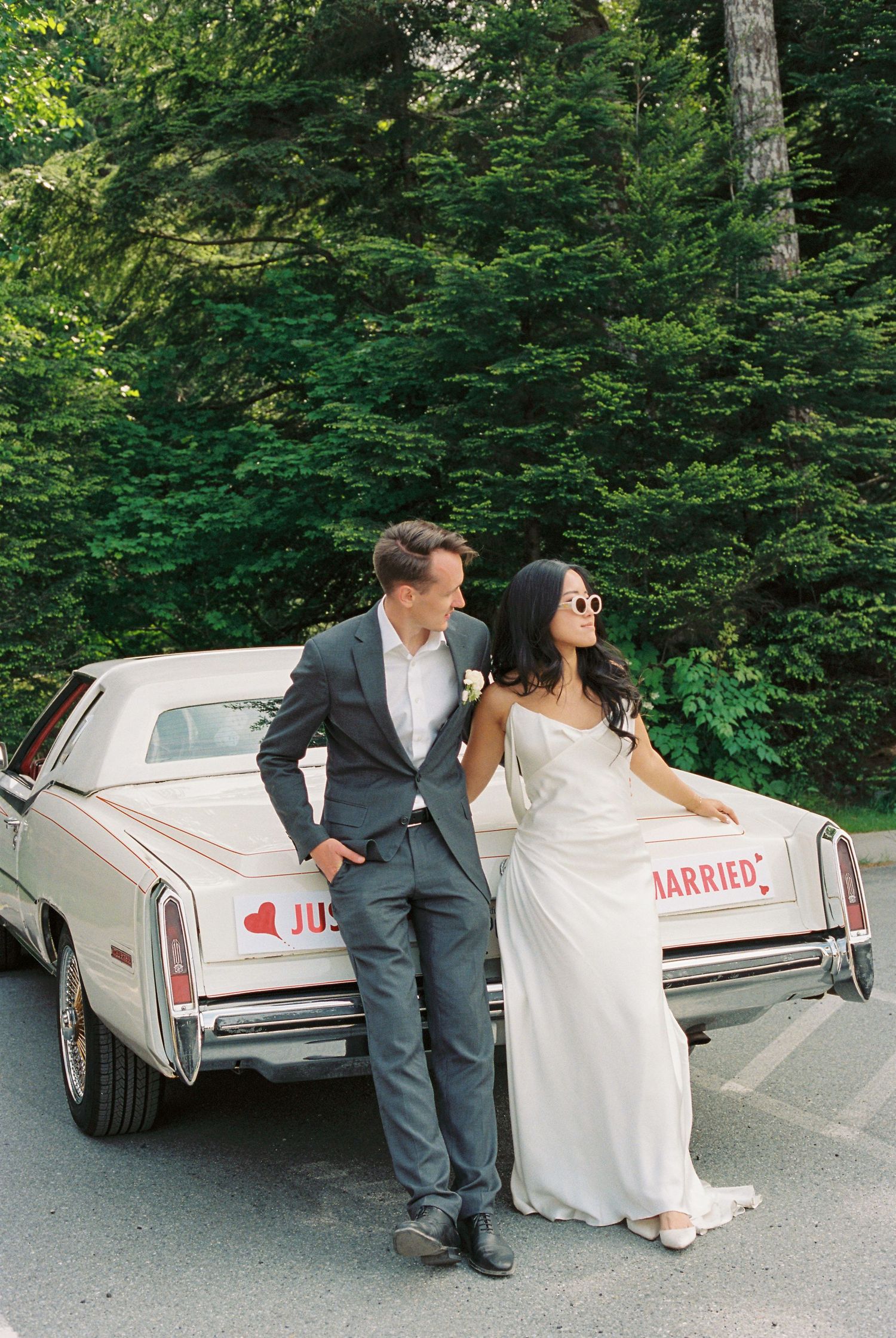 Bride and groom leaning against a vintage Just Married car surrounded by lush green trees on their wedding day.