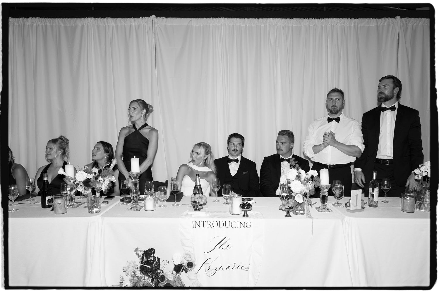 Black and white wedding reception photo of bridal party seated at head table with floral arrangements.