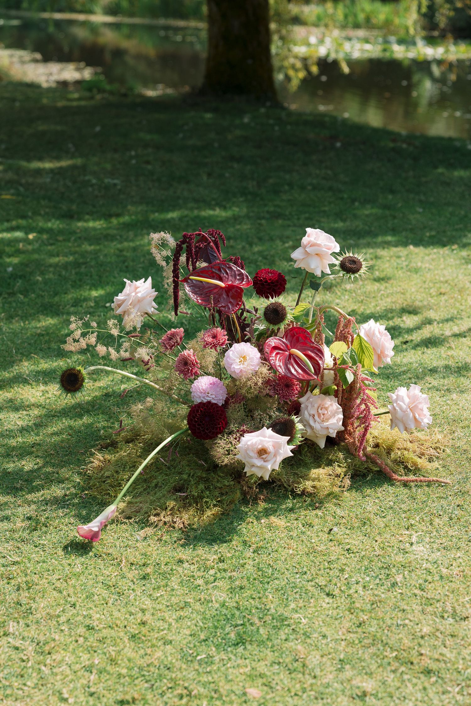 Lush floral arrangement of pink roses, red anthuriums, and dahlias on green grass near a serene garden pond.
