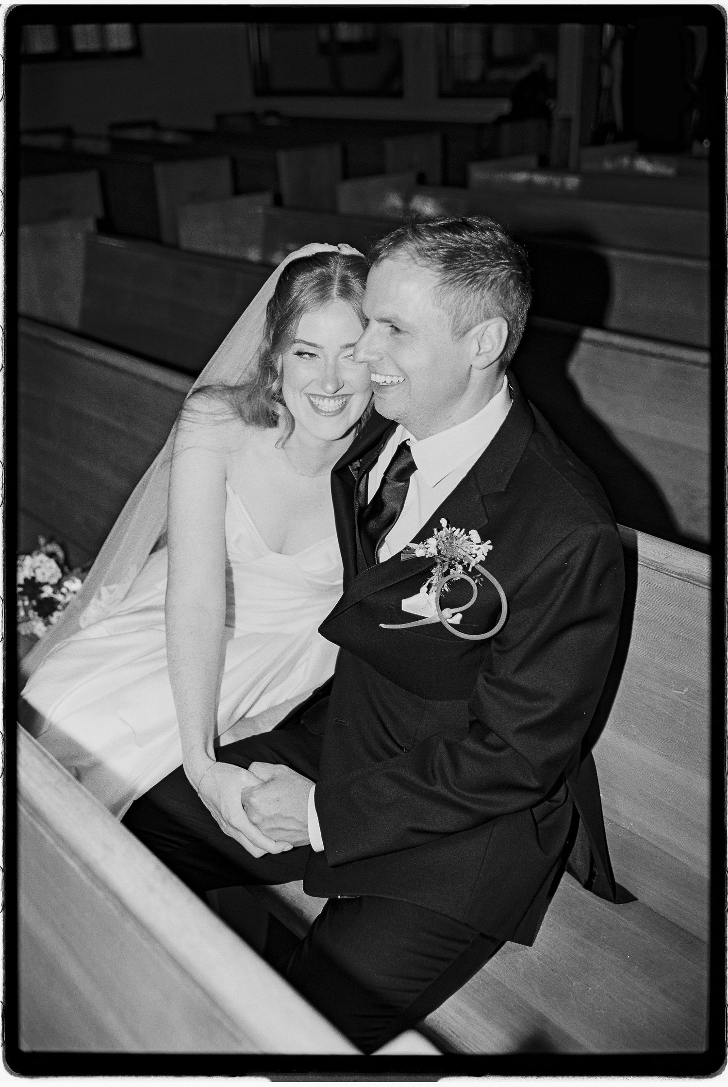 Black and white wedding photo of smiling bride in veil and groom in suit with boutonniere sitting together in church pews.