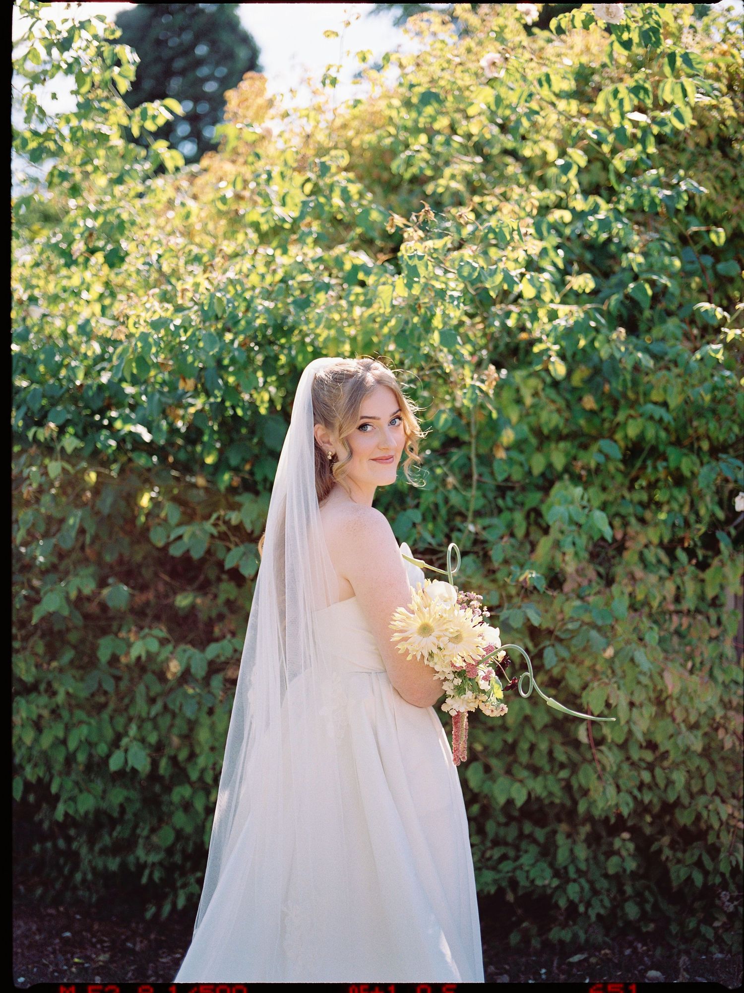Bride in white gown and veil holding floral bouquet, posing outdoors among lush green foliage in soft natural light.