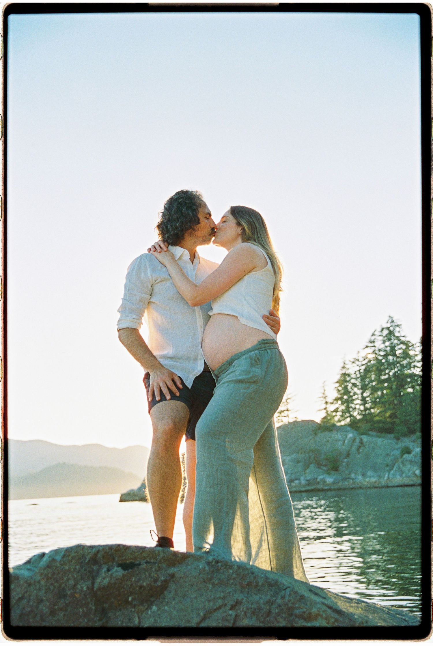 Couple kissing on rocks by the water at sunset, pregnant woman with exposed belly, trees and mountains in background.