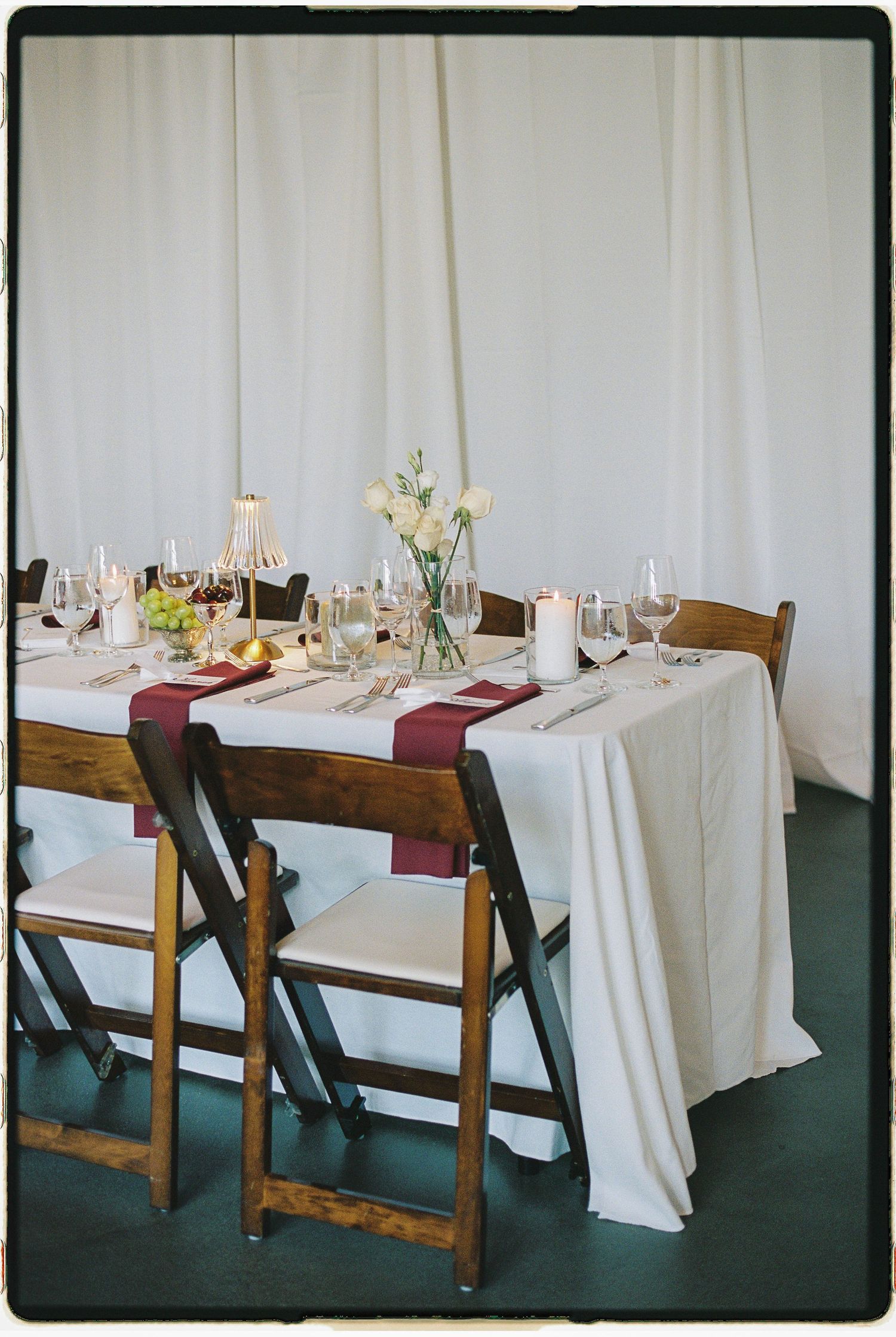 Elegant wedding reception table with white linen, burgundy runners, floral centerpiece, wine glasses, and wooden folding chairs.