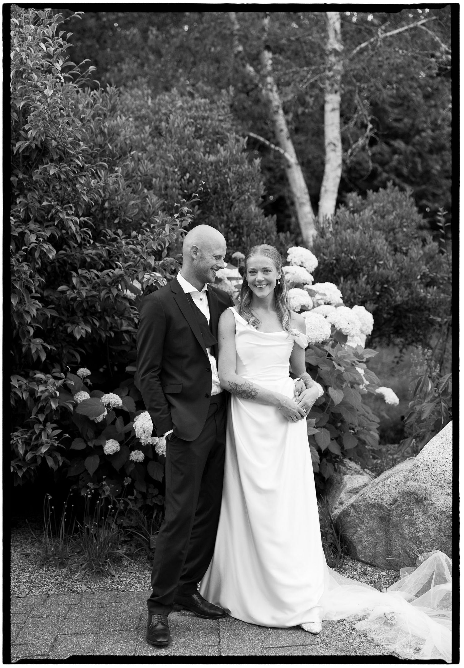 Black and white wedding photo of a bald groom in a suit and smiling bride in a white gown posing in a garden.