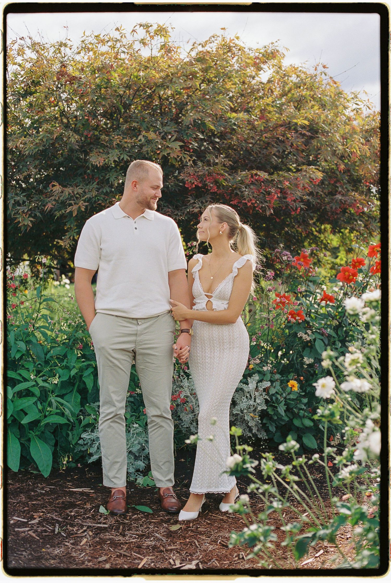 A couple shares a tender moment in a garden filled with blooming red and white flowers.