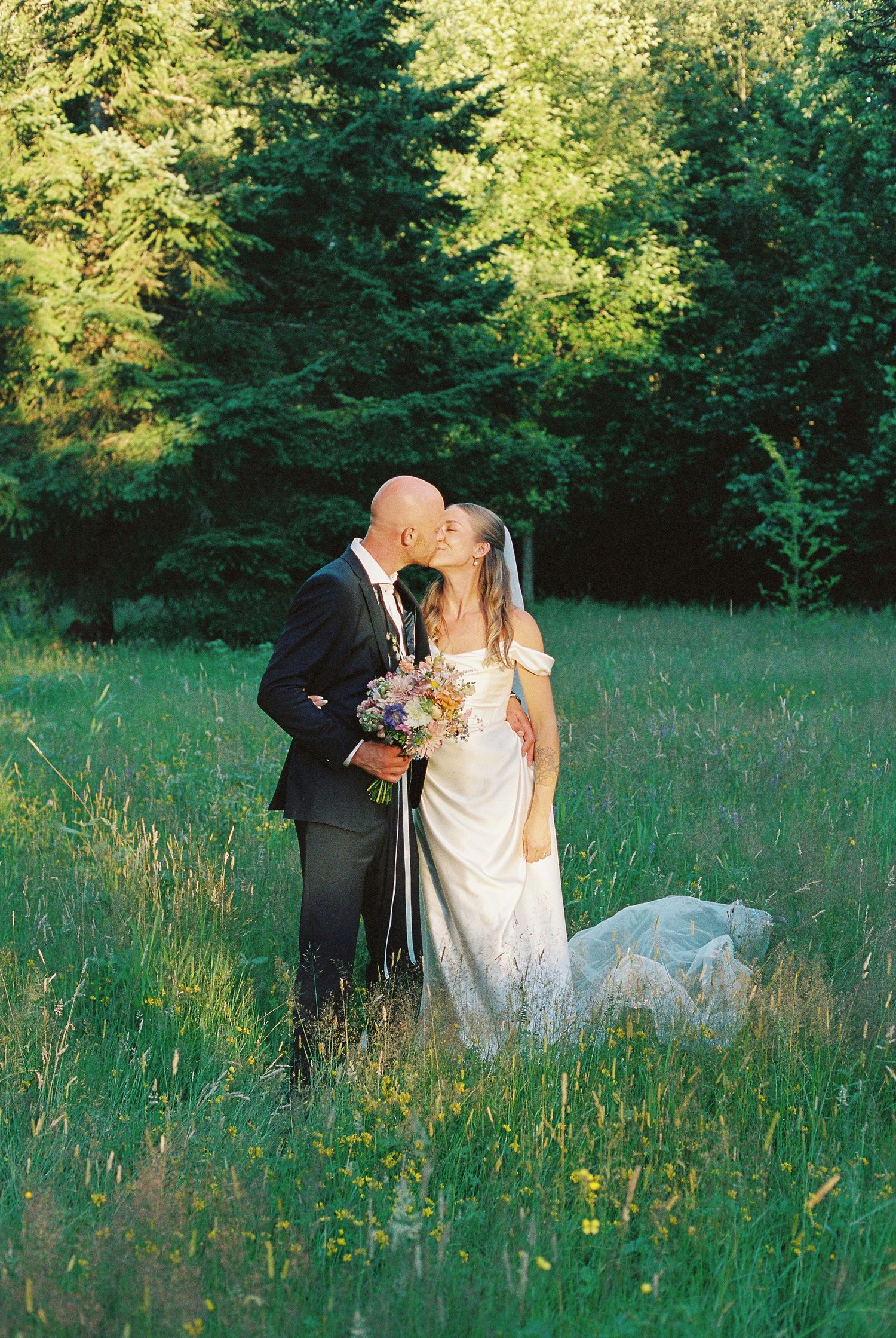 A romantic wedding couple embraces in a sunlit meadow surrounded by evergreen trees.
