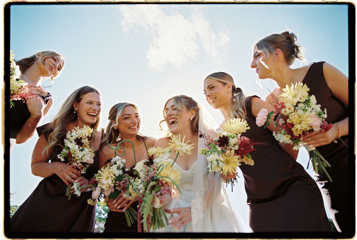 Bride in white gown laughing with bridesmaids in dark dresses holding floral bouquets outdoors on a sunny day.