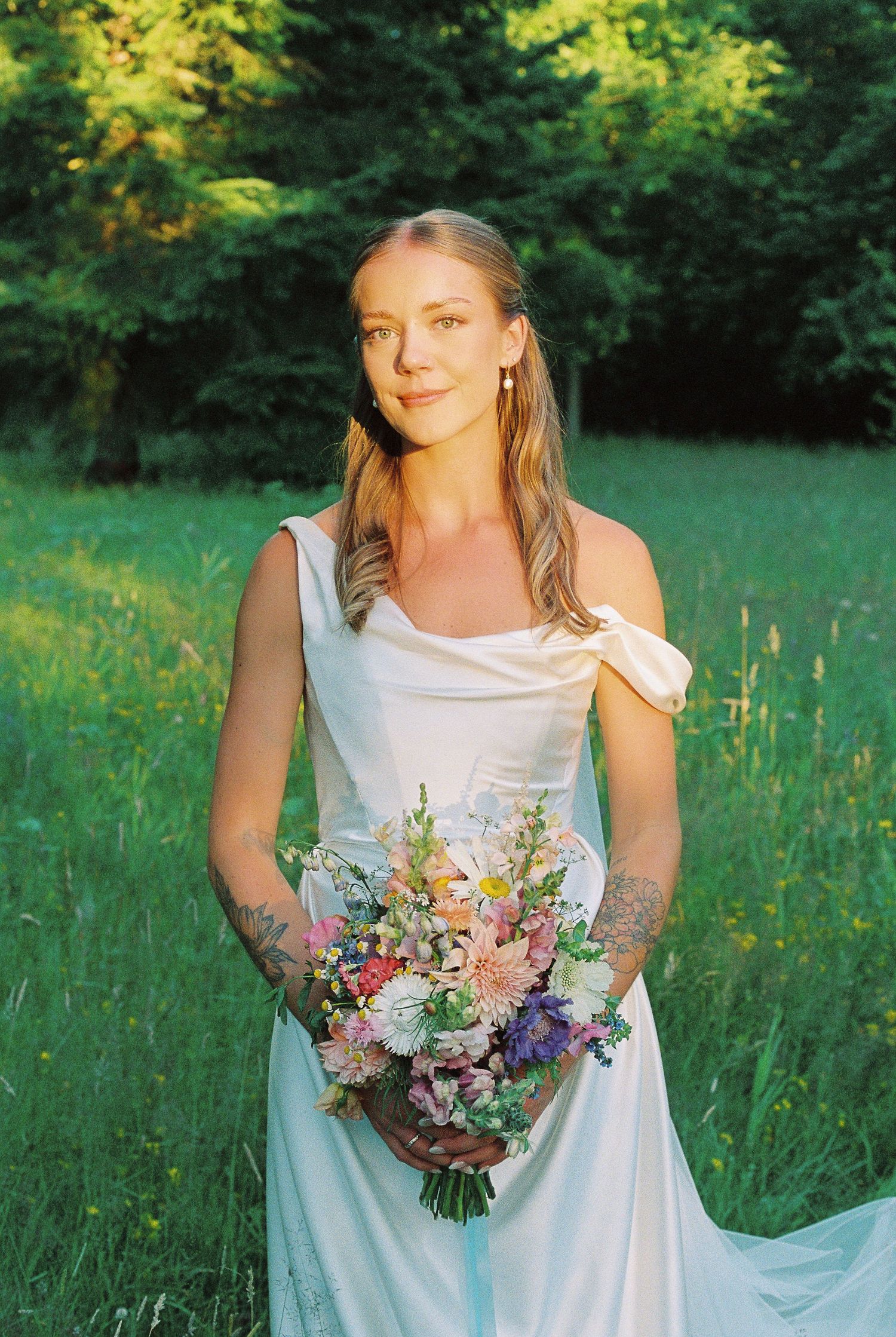A bride in an elegant white dress holds a colorful summer bouquet in a green meadow.