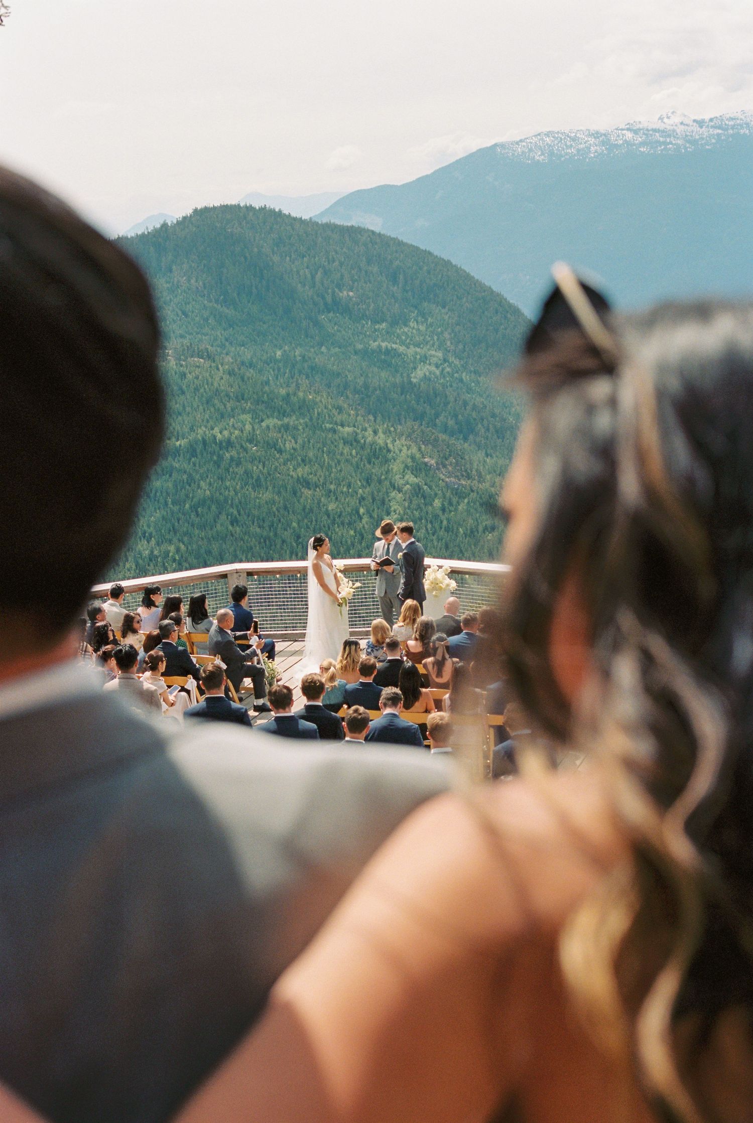 Outdoor mountain wedding ceremony with guests seated overlooking lush green peaks and snow-capped mountains under a clear sky.