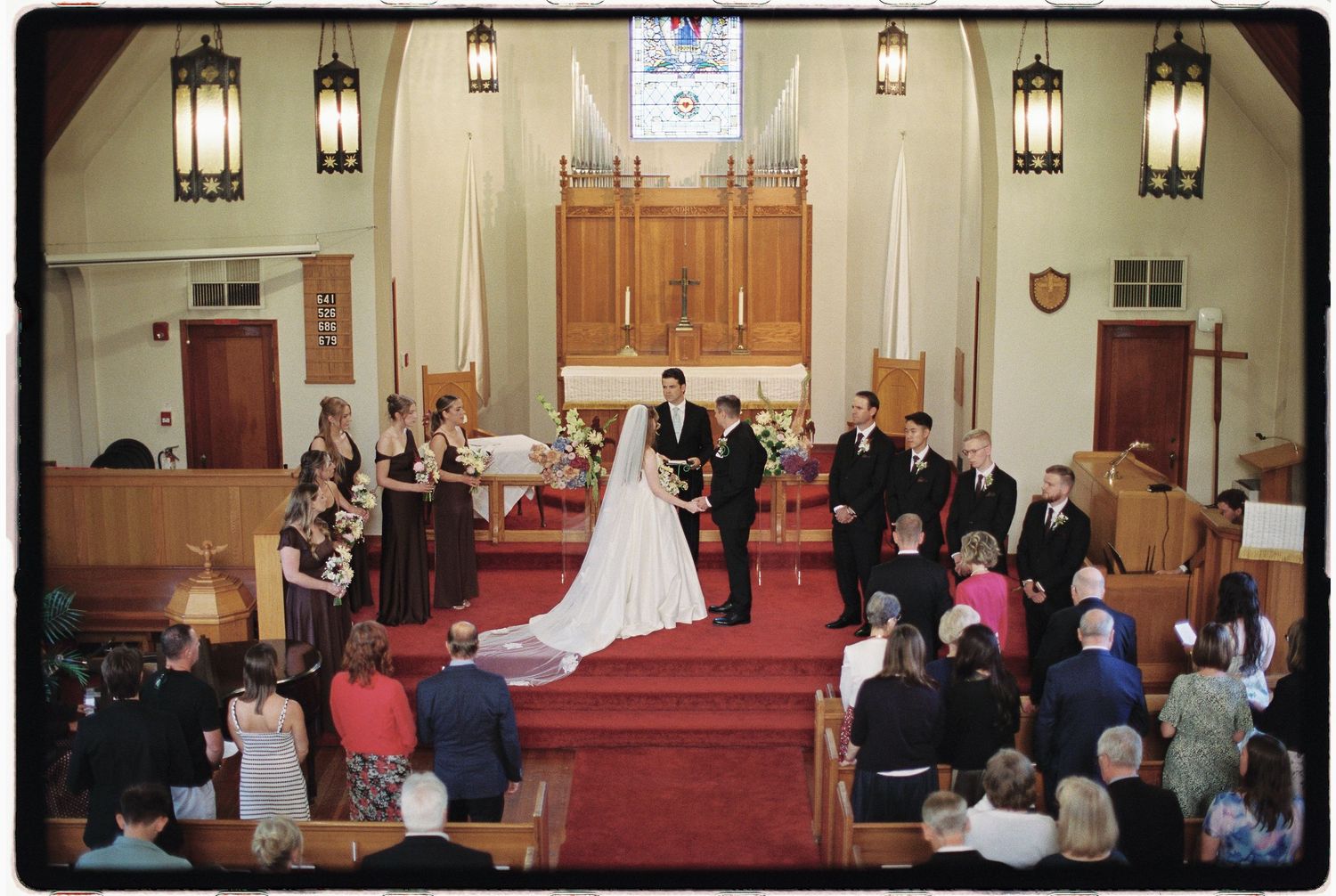 Elegant church wedding ceremony with bride and groom at altar, guests seated in wooden pews, red carpet aisle, and stained glass windows.