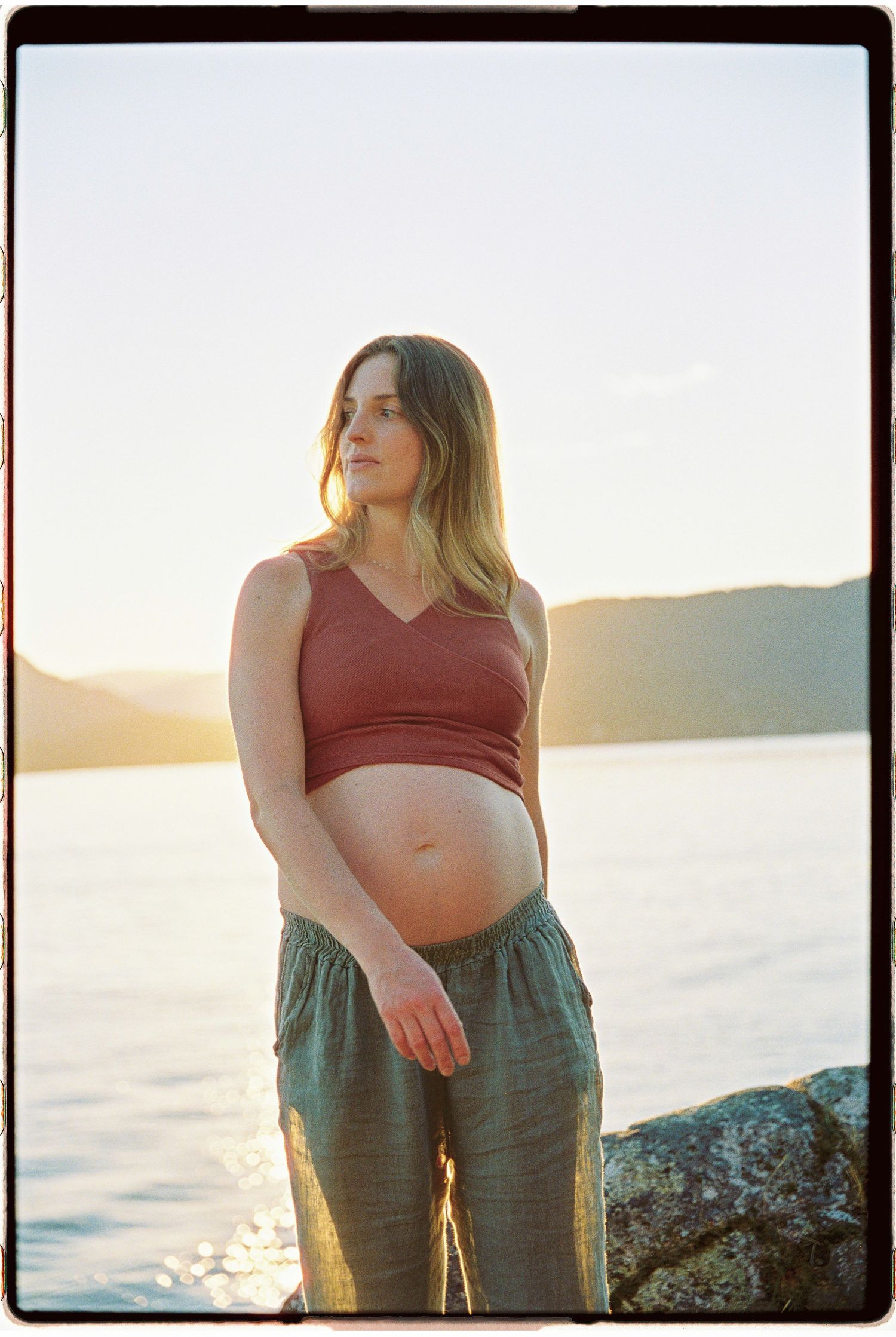 Pregnant woman in rust crop top and teal pants stands on rocks by a lake at golden hour sunset.