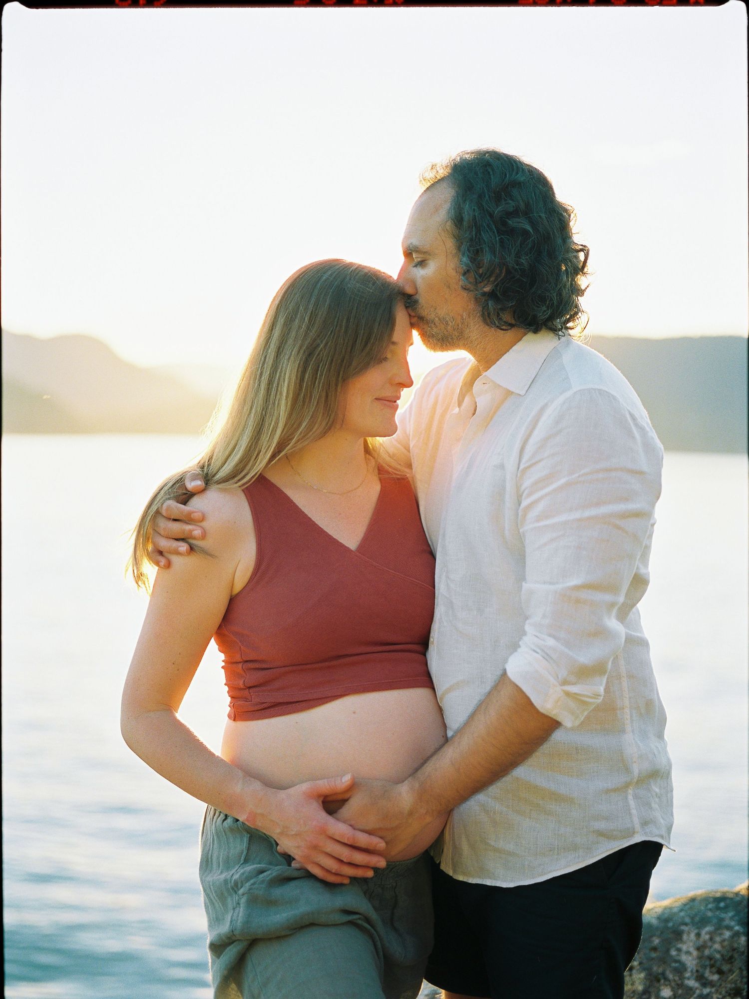 Couple sharing a tender moment at sunset by the water, pregnant woman in red crop top cradled by partner.