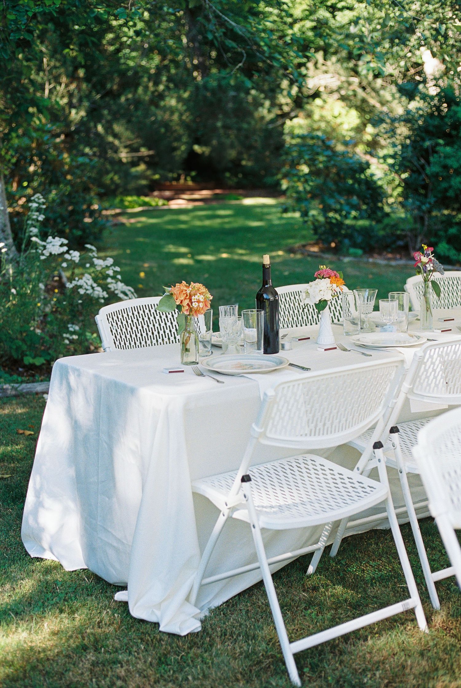 Elegant outdoor garden dining table with white linen, folding chairs, orange floral centerpieces, and a wine bottle on green lawn.