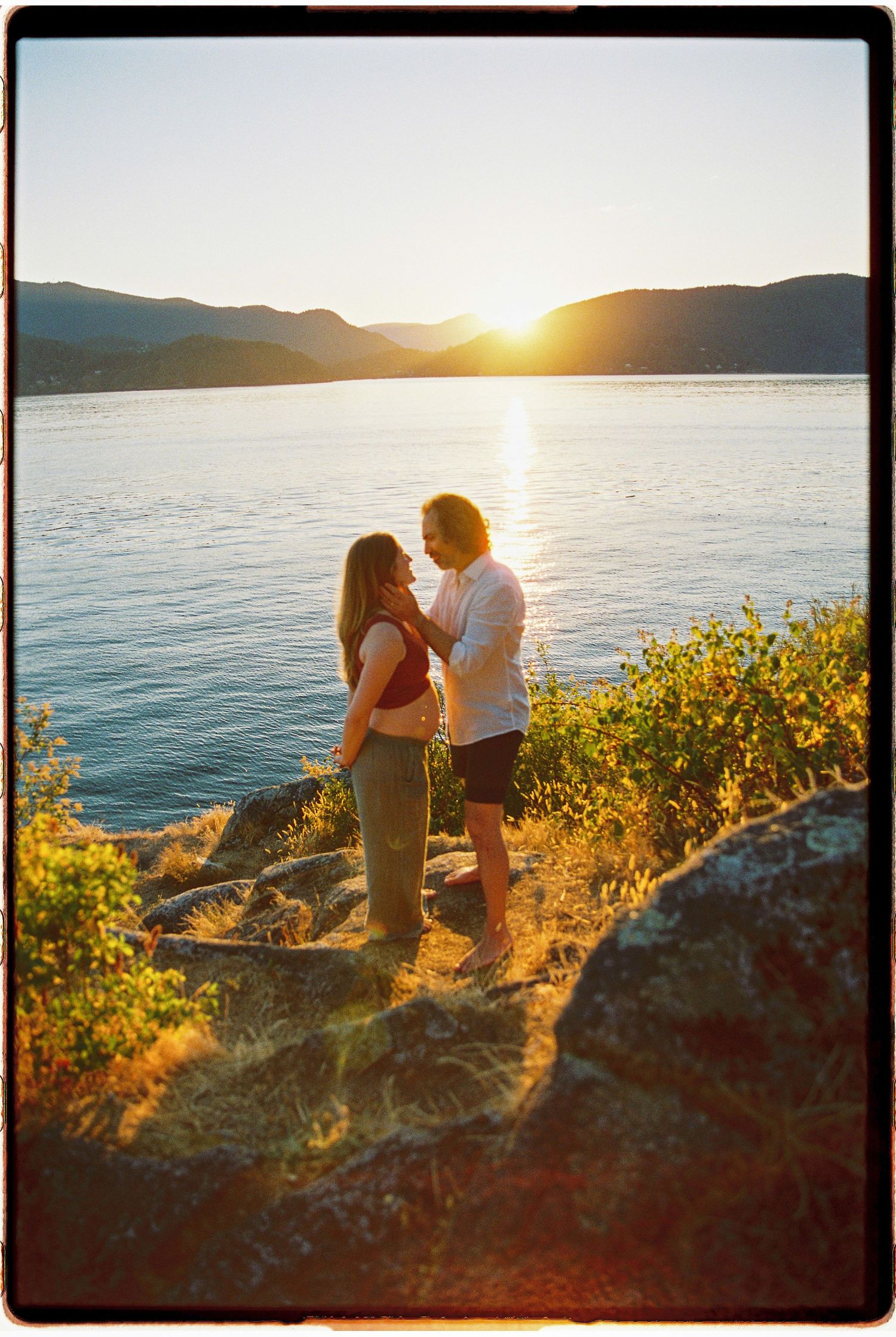 Couple embracing at golden hour sunset by a mountain lake, standing on rocky shoreline with glowing sun behind hills.