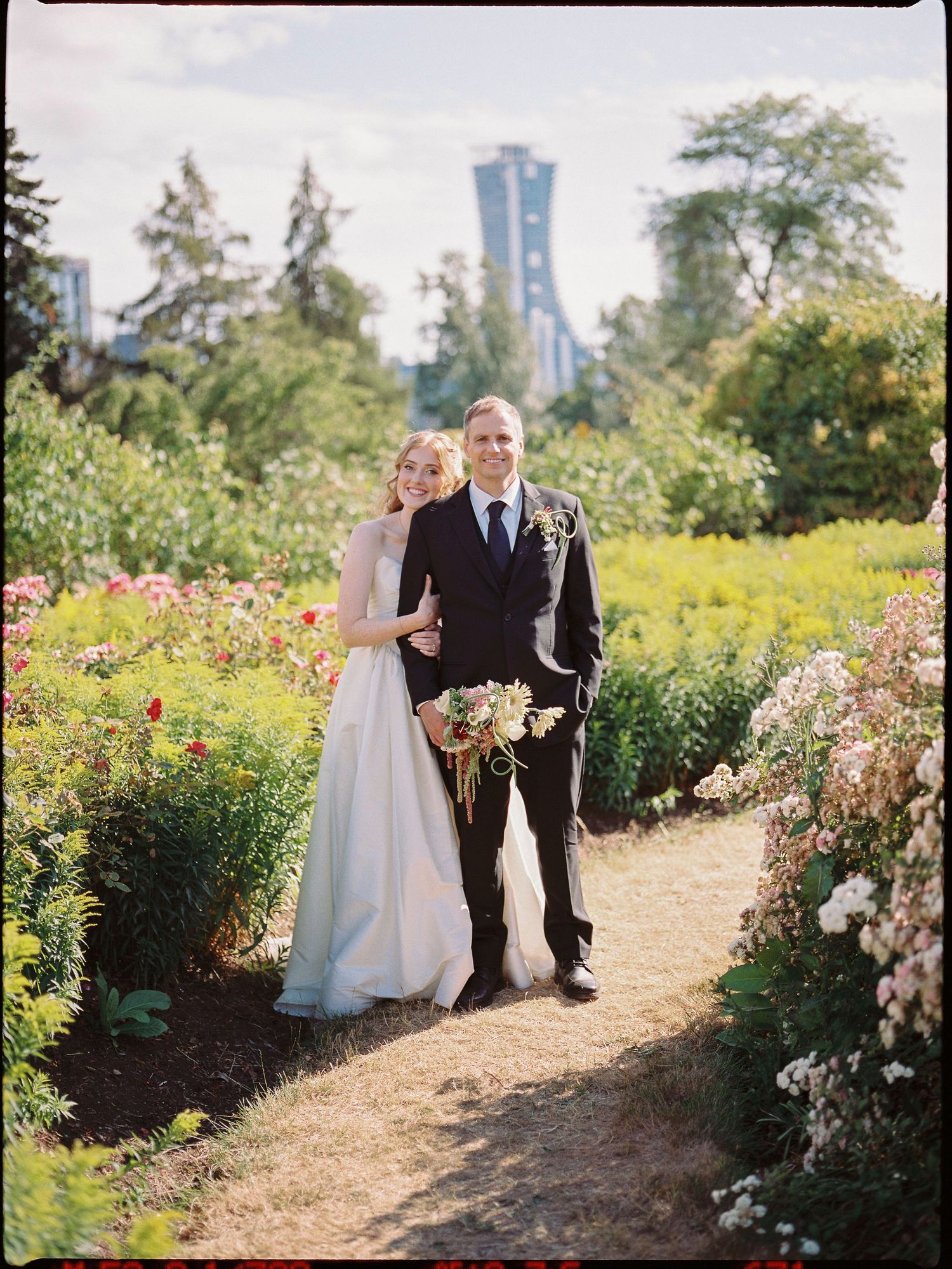 Bride and groom posing on a garden path surrounded by colorful flowers with city skyline in background.